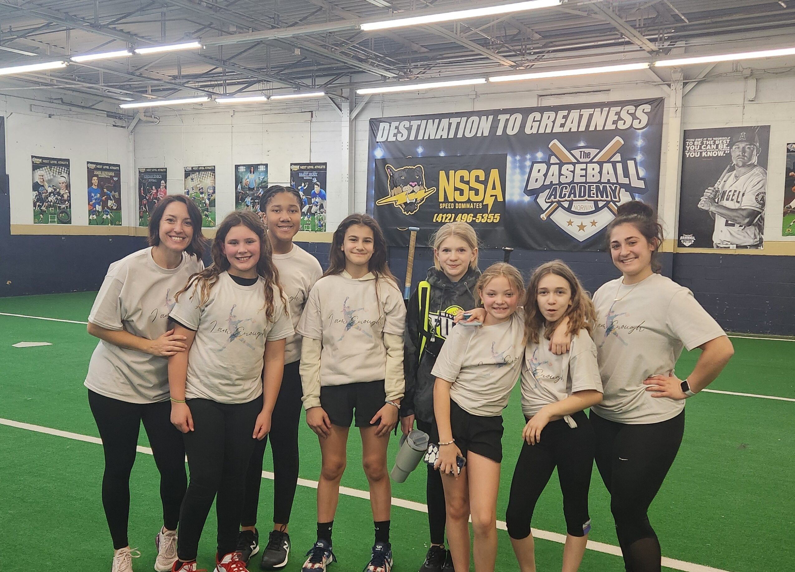 Group of eight girls standing on an indoor sports field, smiling, with banners and sports equipment in background.