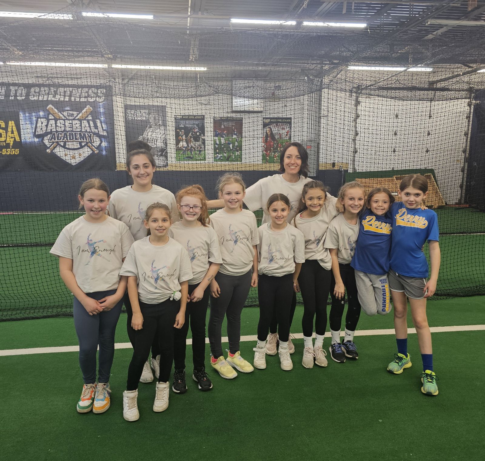 Group of ten children standing on an indoor sports field, smiling, with a netted background and sports banners.