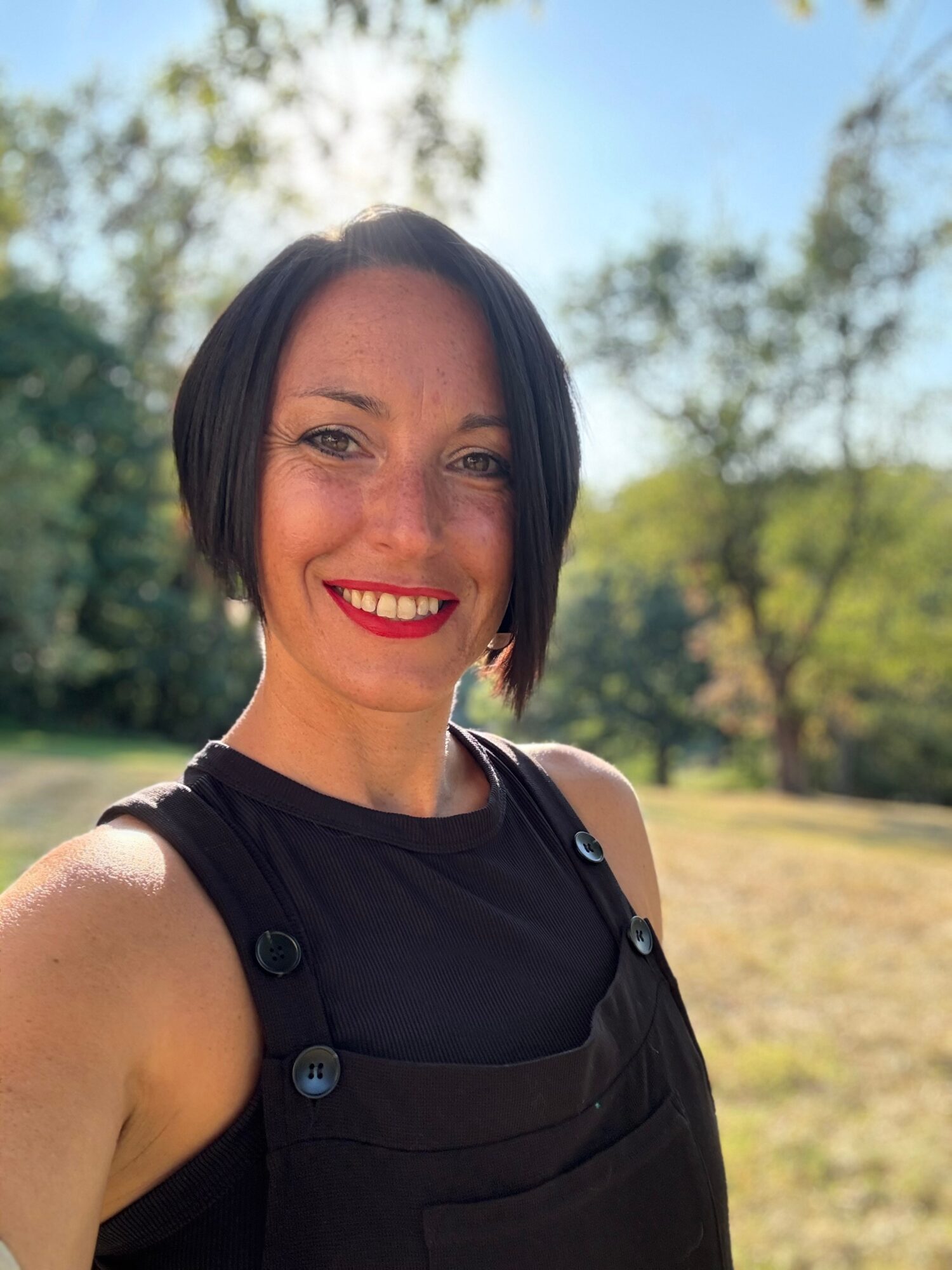 Smiling woman with short dark hair outdoors, wearing a black sleeveless top, trees and sky in background.