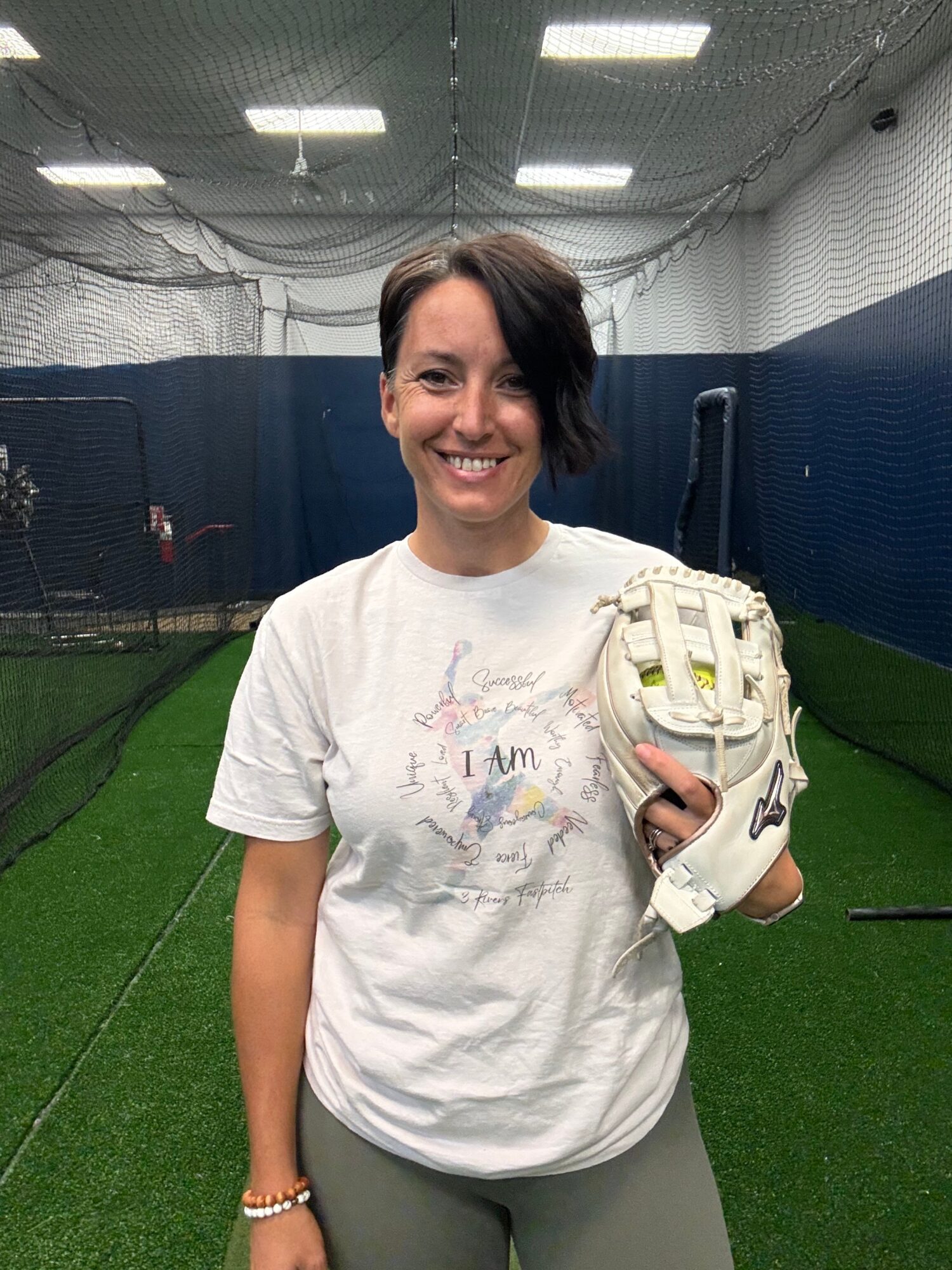Woman smiling in indoor sports facility holding a baseball glove, wearing a white T-shirt and gray pants.