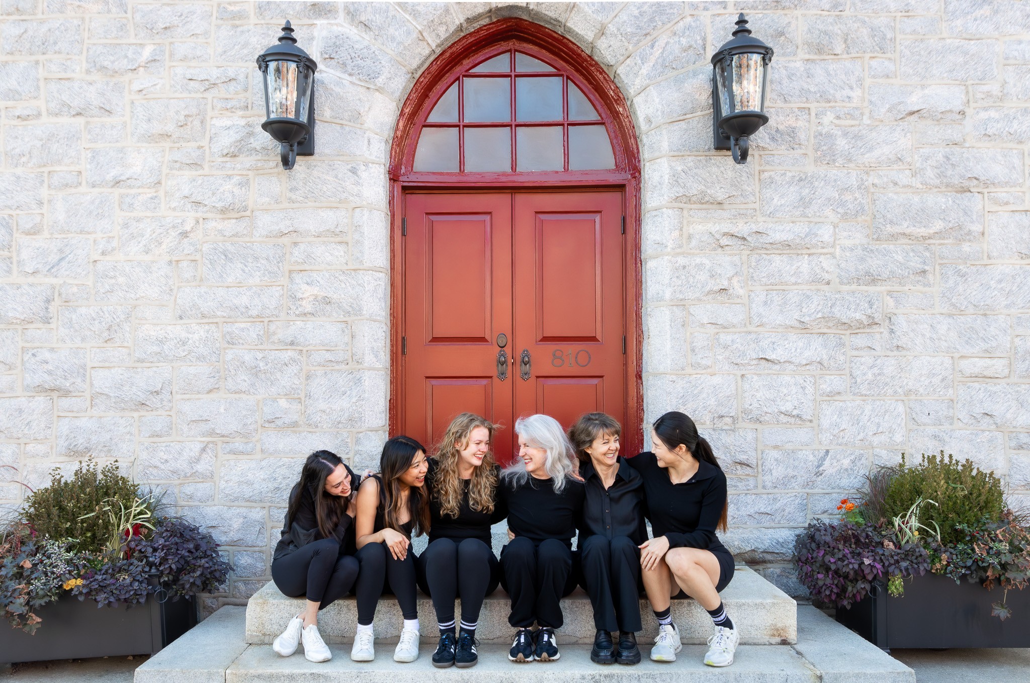 Group of six people sitting on steps in front of a red arched door with stone wall background.