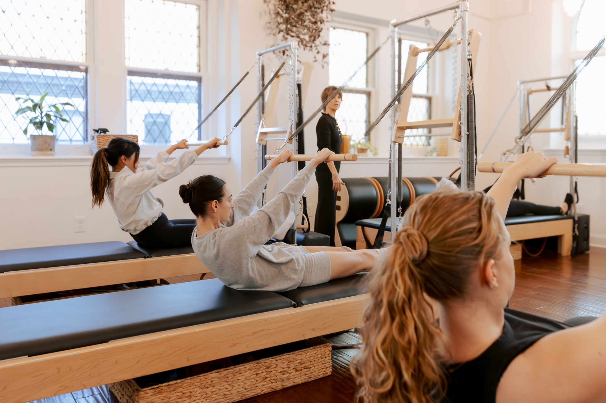 Group of women exercising with resistance bands in a fitness studio, some lying on mats and others standing, with large windows in background.