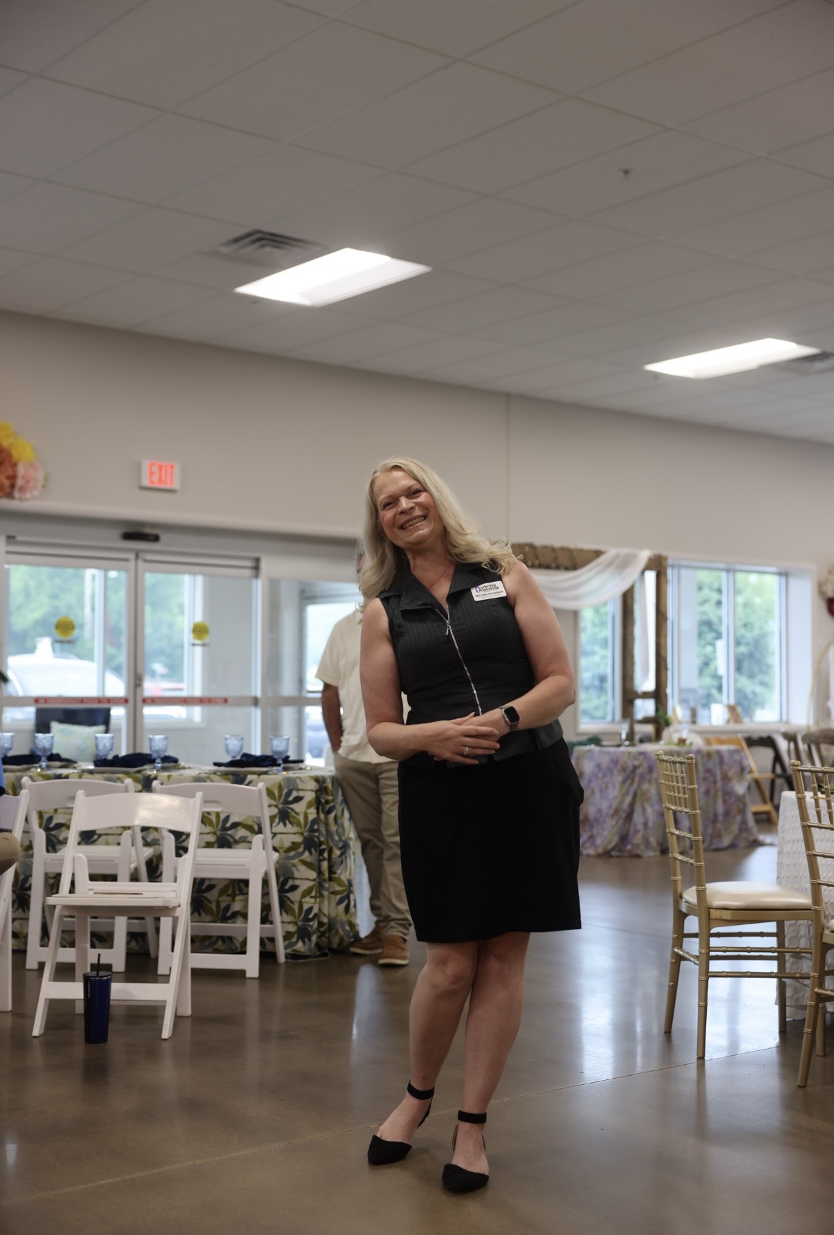 Woman in black dress smiling in a decorated event space with tables and chairs, windows in the background.