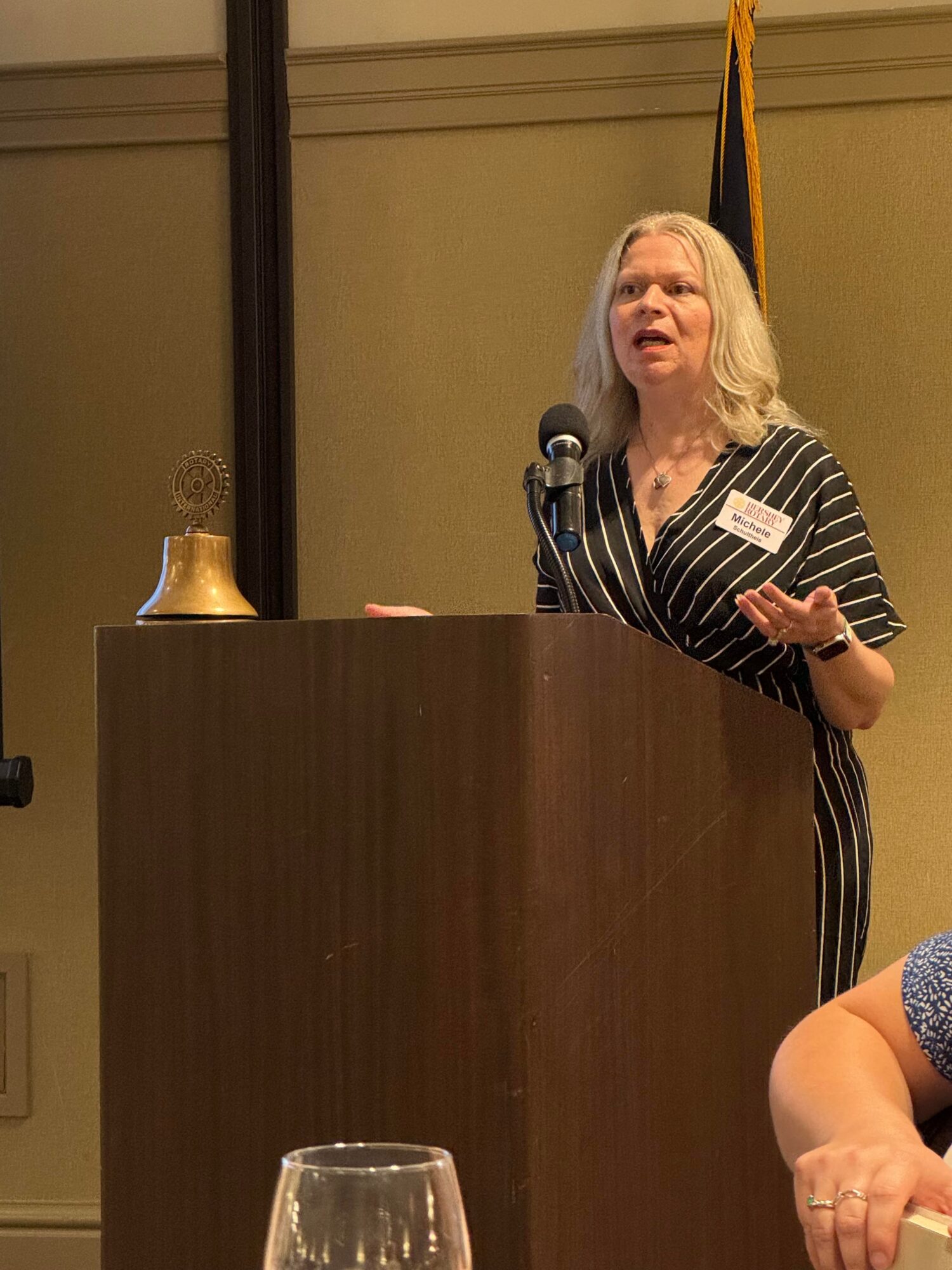 Woman with long gray hair speaking at a podium, gesturing with her right hand, in a room with a bell and flag behind her.