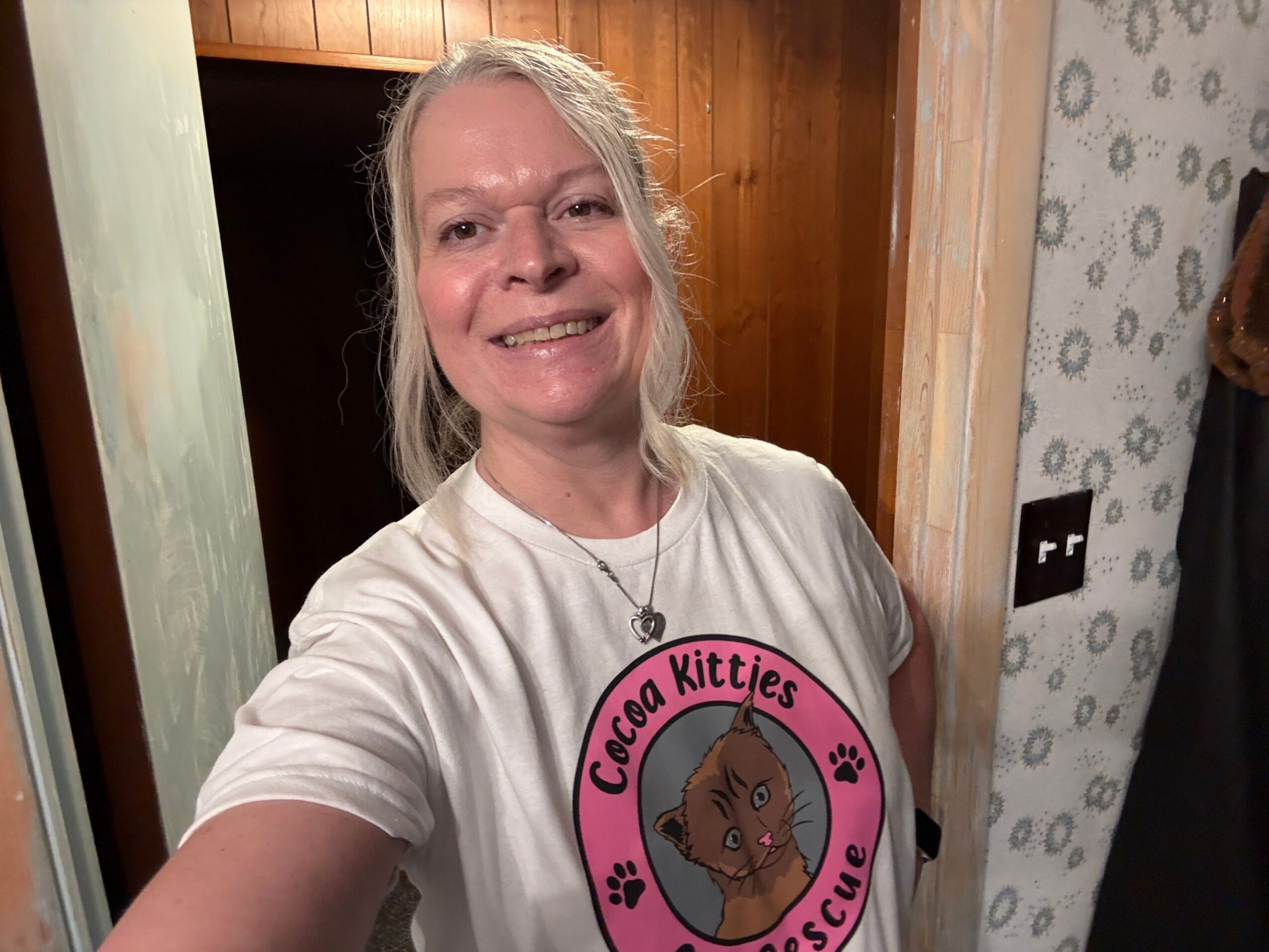 Woman with blonde hair smiling, wearing a white shirt with a pink circular sticker, in a room with wooden and glass surfaces.