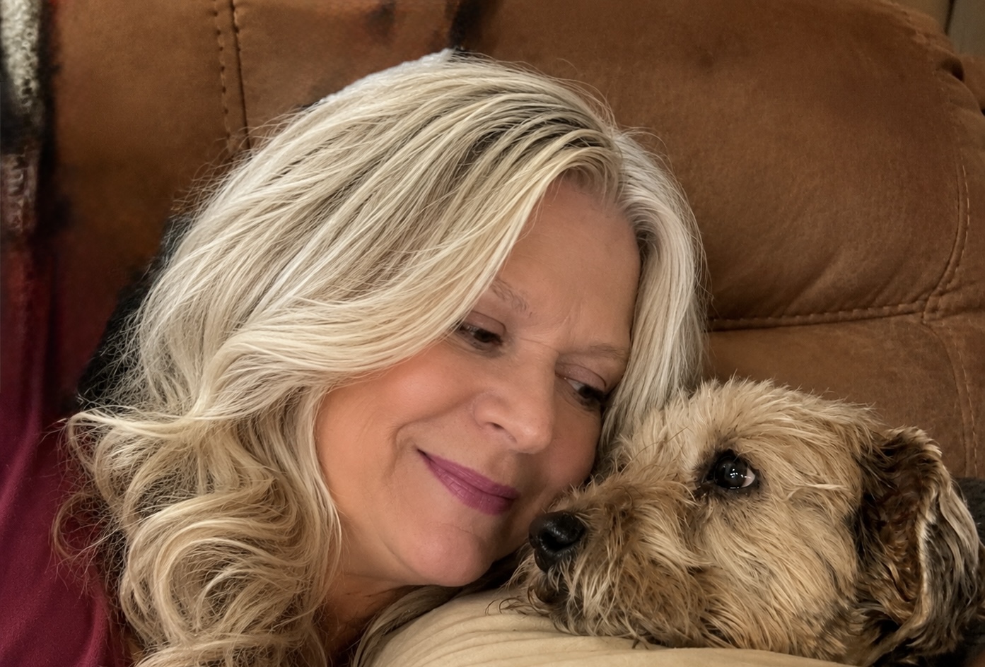 Woman with blonde hair lying on a couch, cuddling a small dog, both appear relaxed and content.