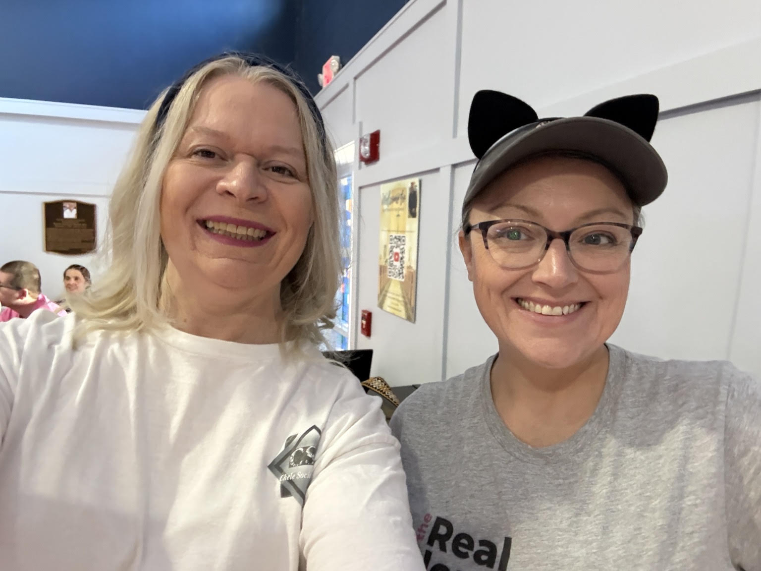Two women smiling, one wearing a white shirt and the other wearing a gray shirt with cat ears hat, in an indoor setting.