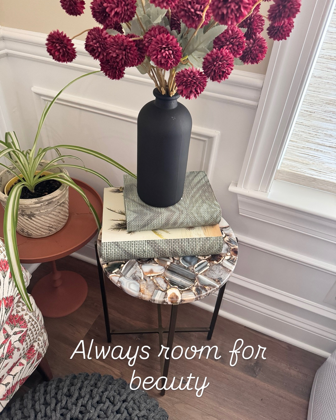 Black vase with purple flowers on a stack of books on a small round table, near a window, with a plant nearby. Text reads 'Always room for beauty'.