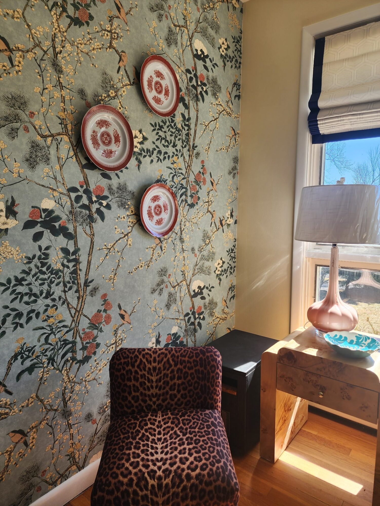 Leopard print chair in front of a floral wallpapered wall with three decorative plates, next to a window with a lamp on a table.