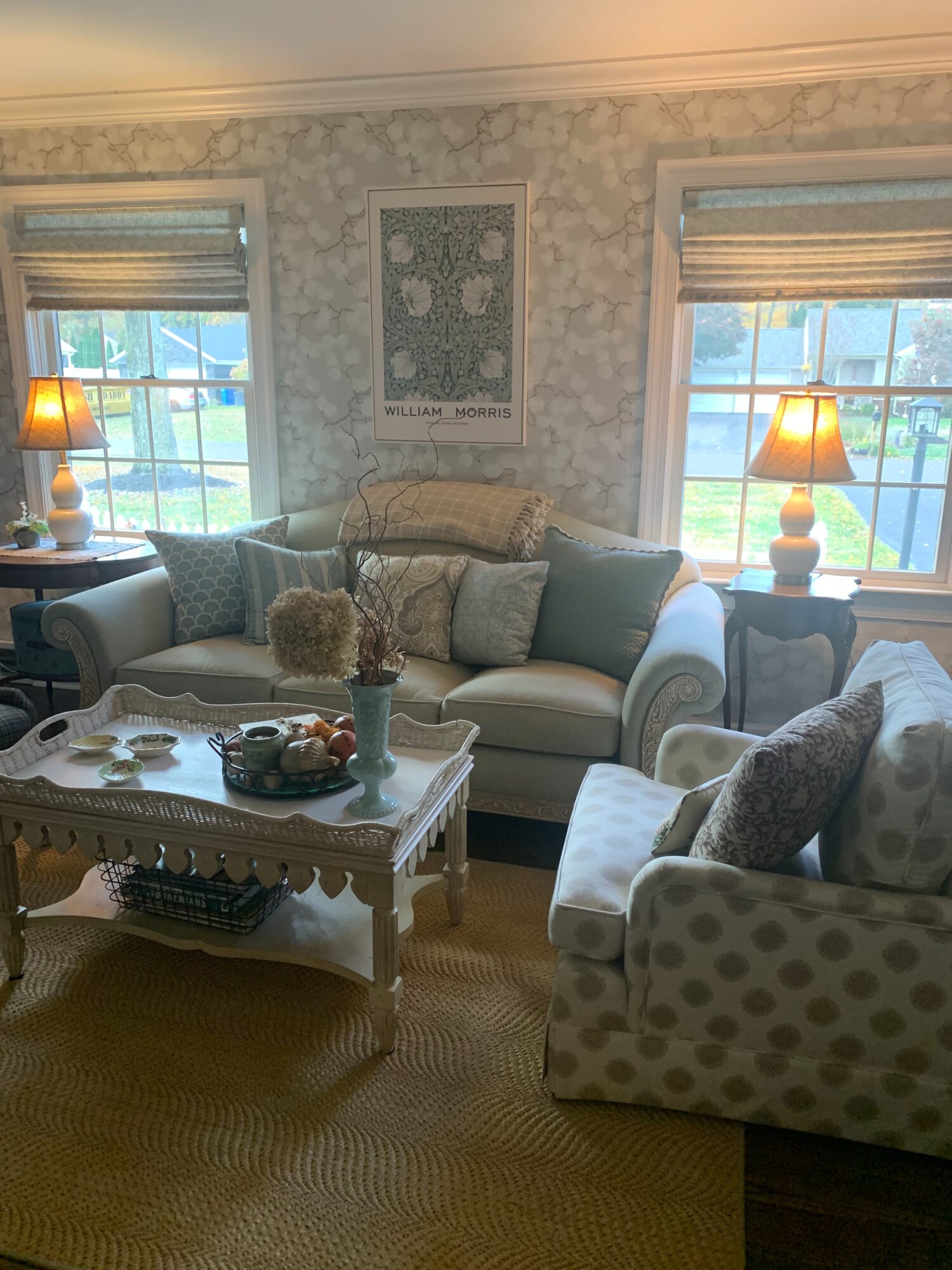 Living room with sofa, armchair, coffee table, two windows with blinds, and lamps on side tables, decorated with patterned wallpaper.