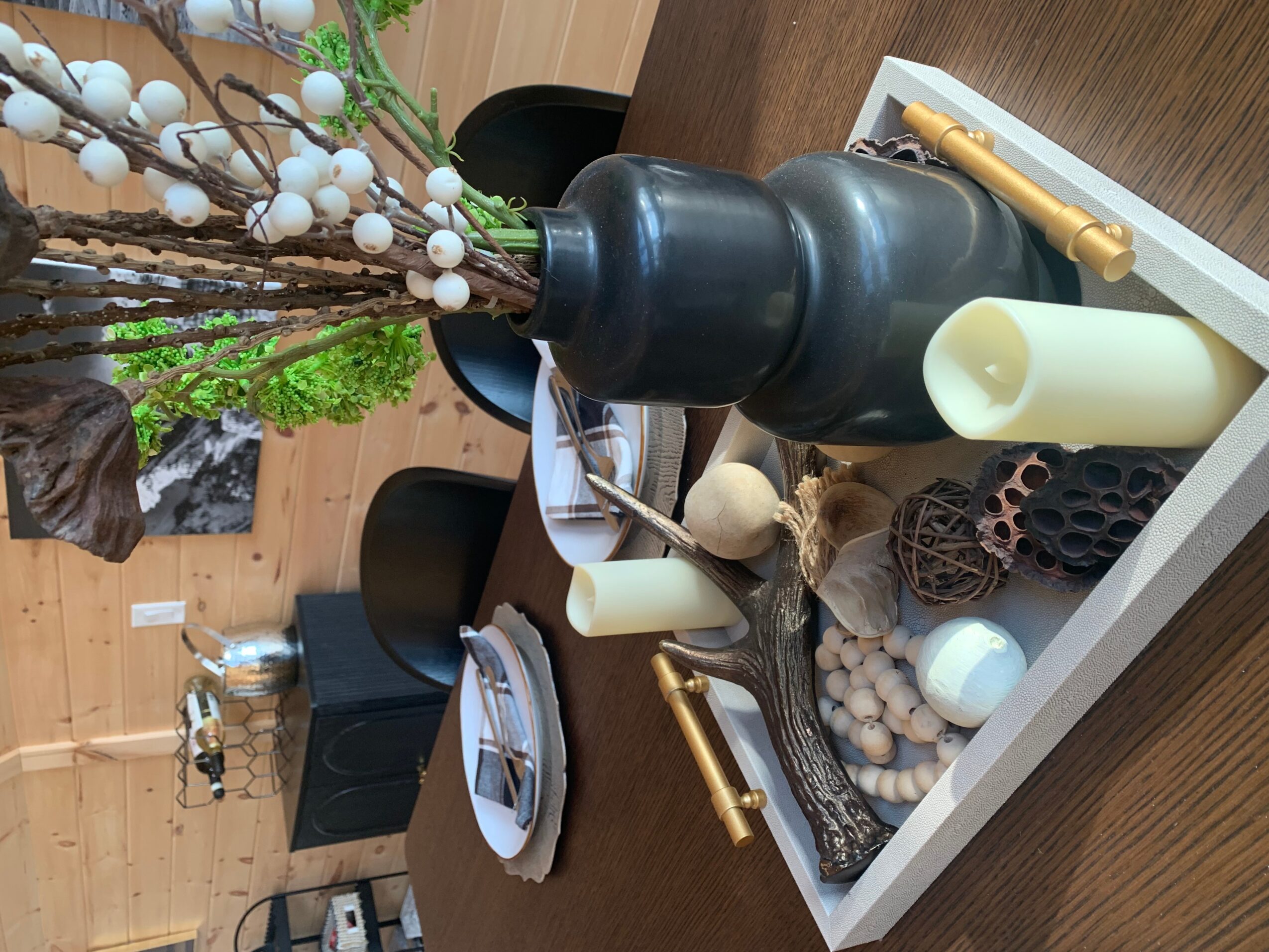 Decorative tray with candles, pinecones, and a black vase with branches and white berries, on a wooden table.