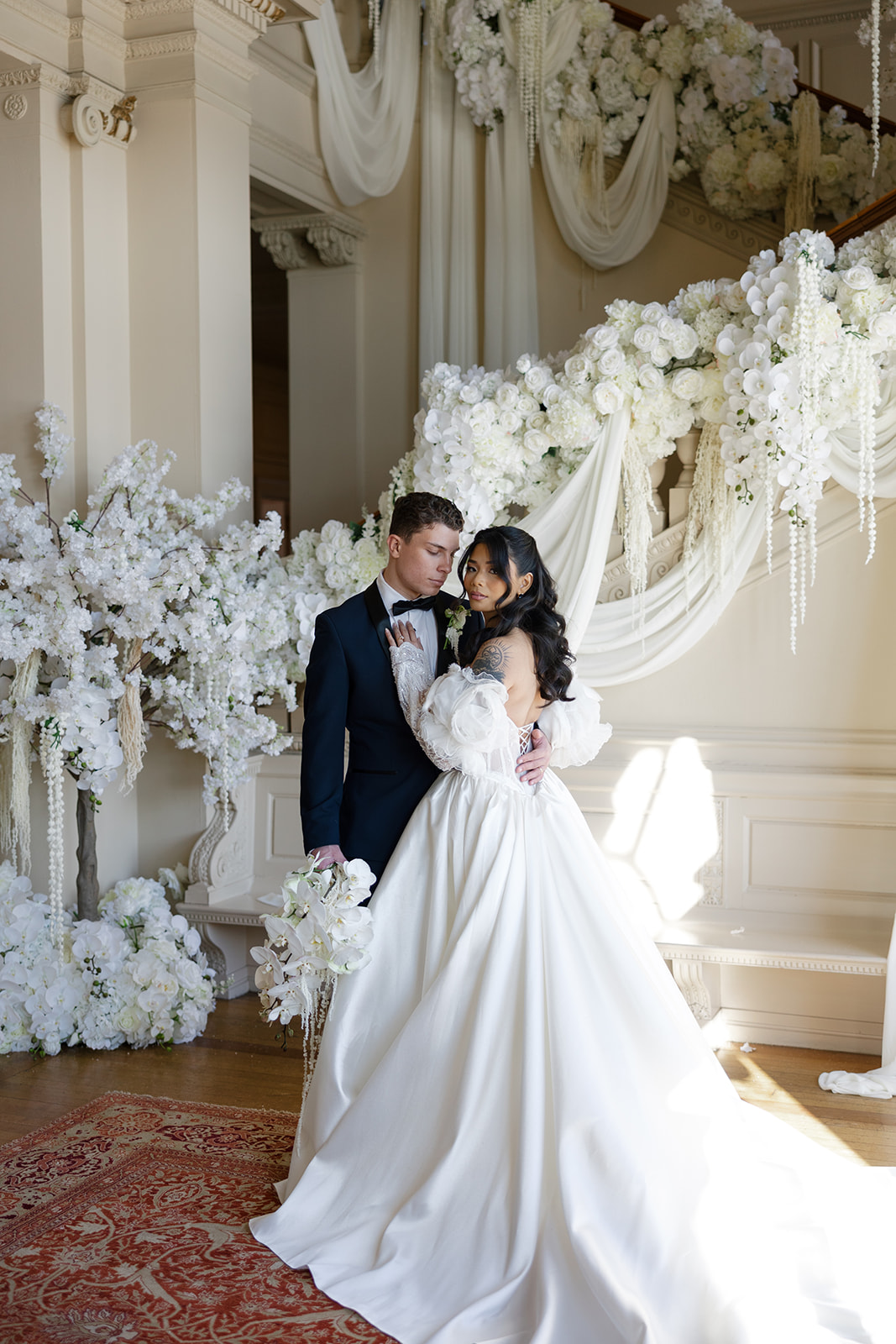 Bride and groom dancing indoors with floral decorations and draped fabric, sunlight streaming in, elegant setting.