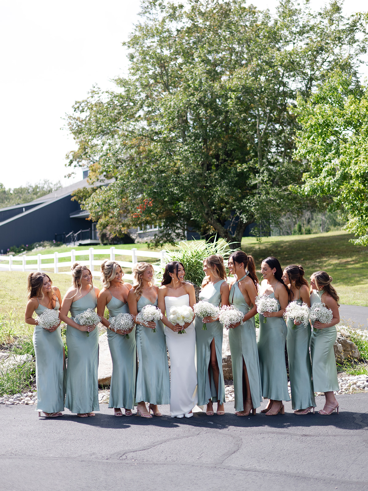 Group of women in light blue dresses holding white bouquets standing outdoors on a sunny day with trees in the background.