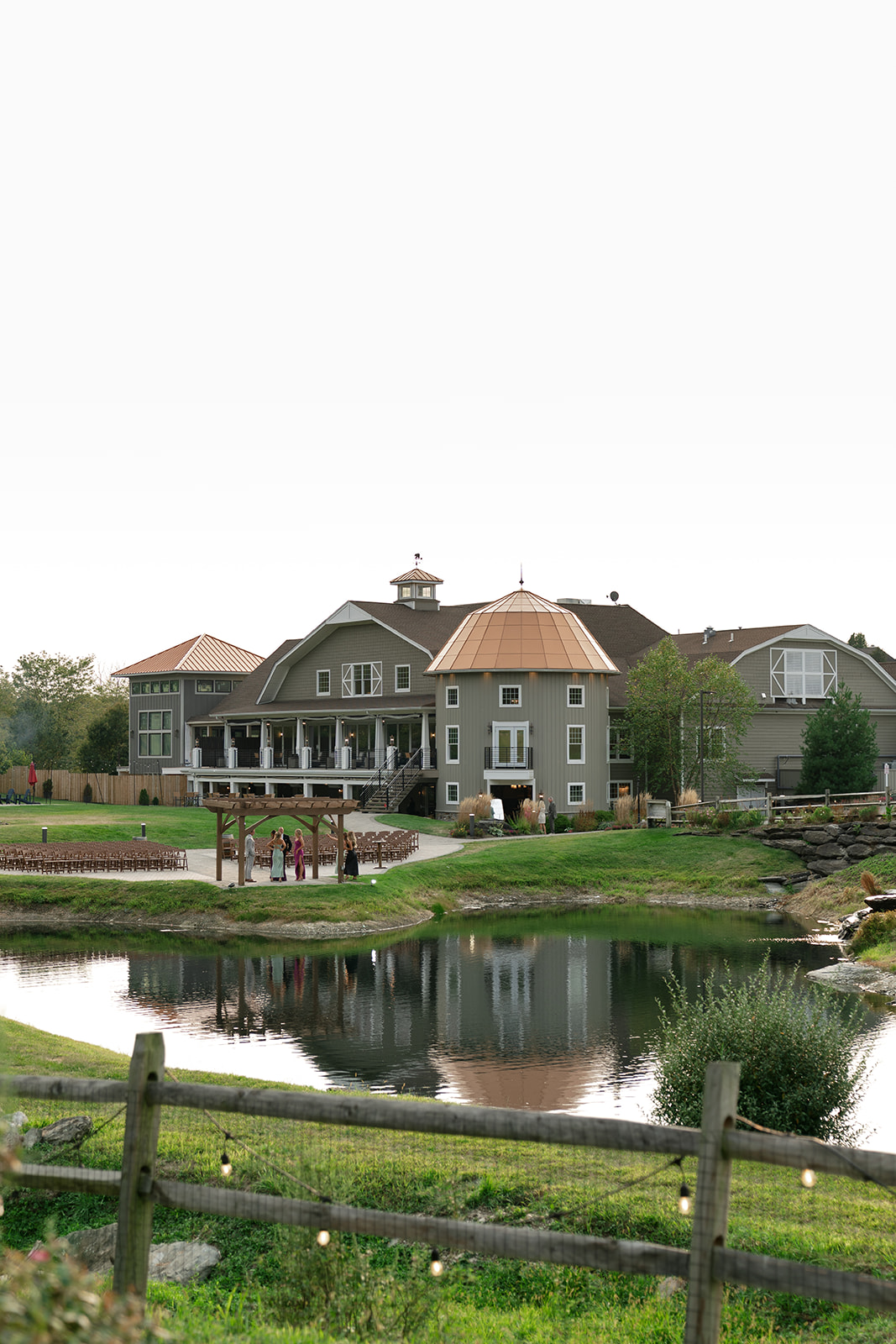 Large house with multiple roofs and a tower, reflected in a pond, surrounded by a grassy area and a wooden fence.