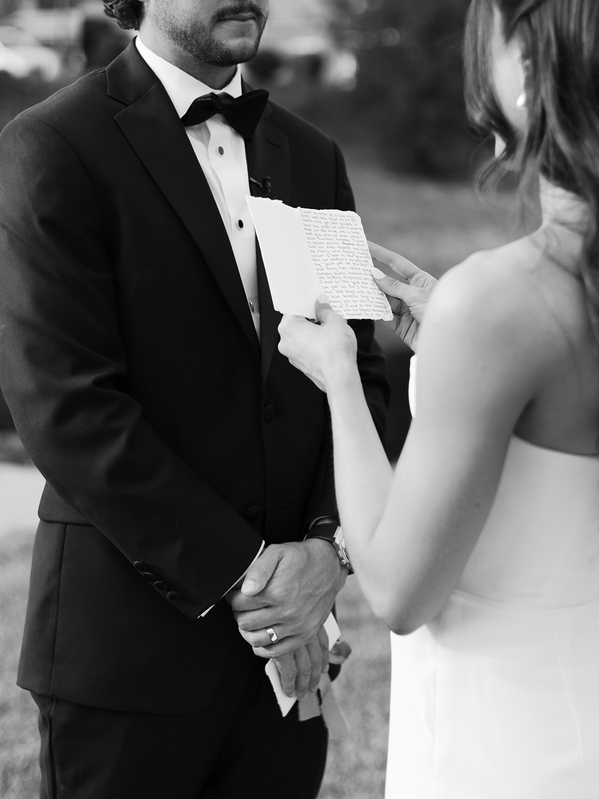 Woman holding a small gift or card in front of a man in a tuxedo outdoors.