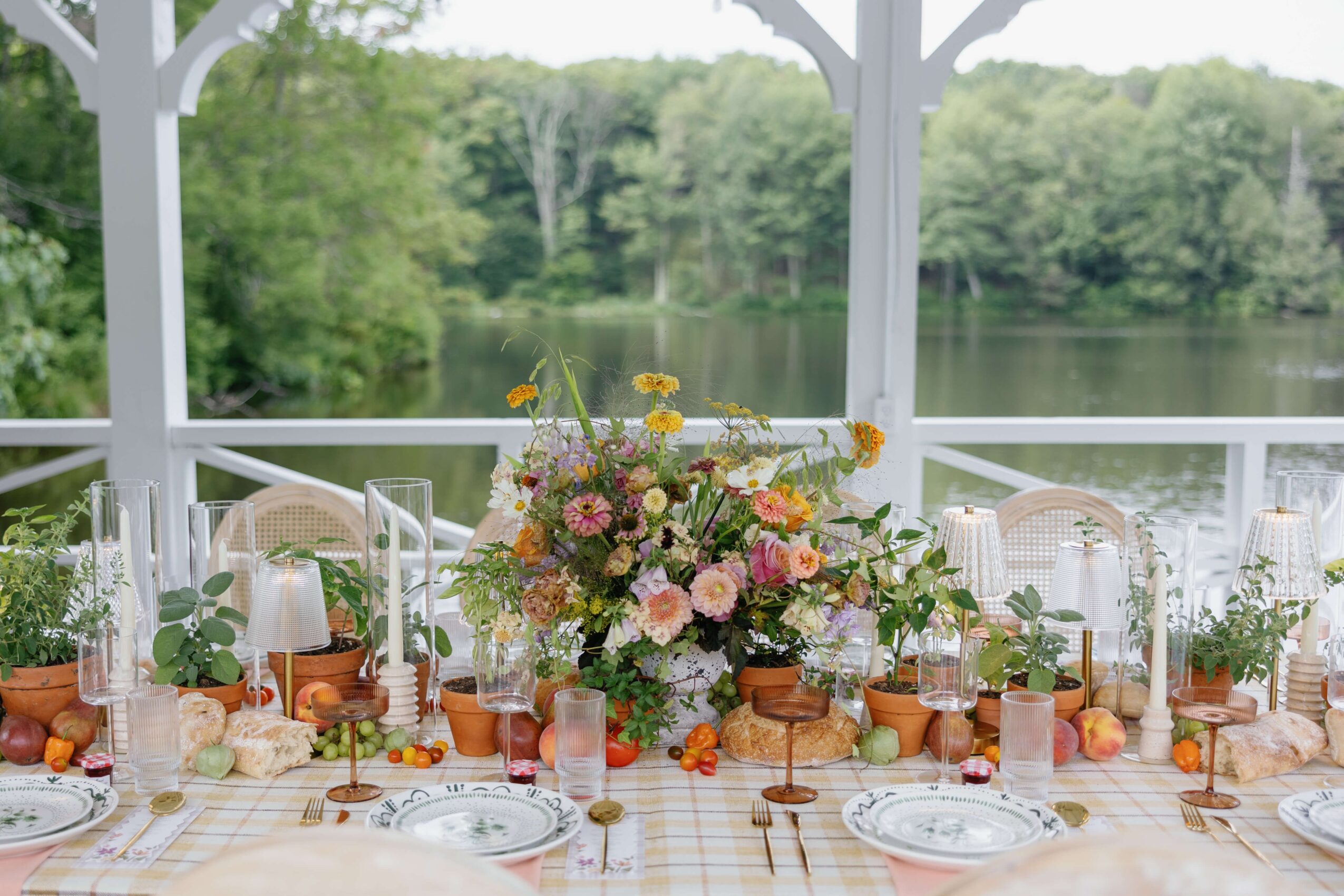 A table set for a meal with a large floral centerpiece, surrounded by candles, plates, and glasses, overlooking a lake and trees.