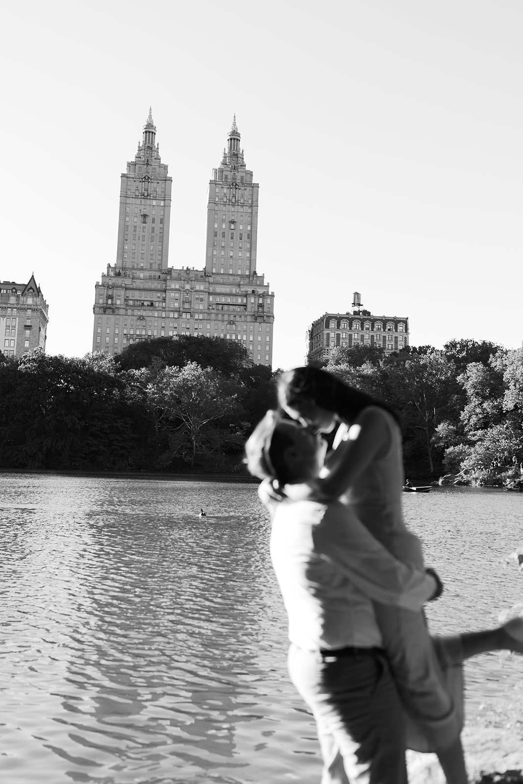 Couple kissing near water with city skyline and tall buildings in background, black and white, romantic scene.