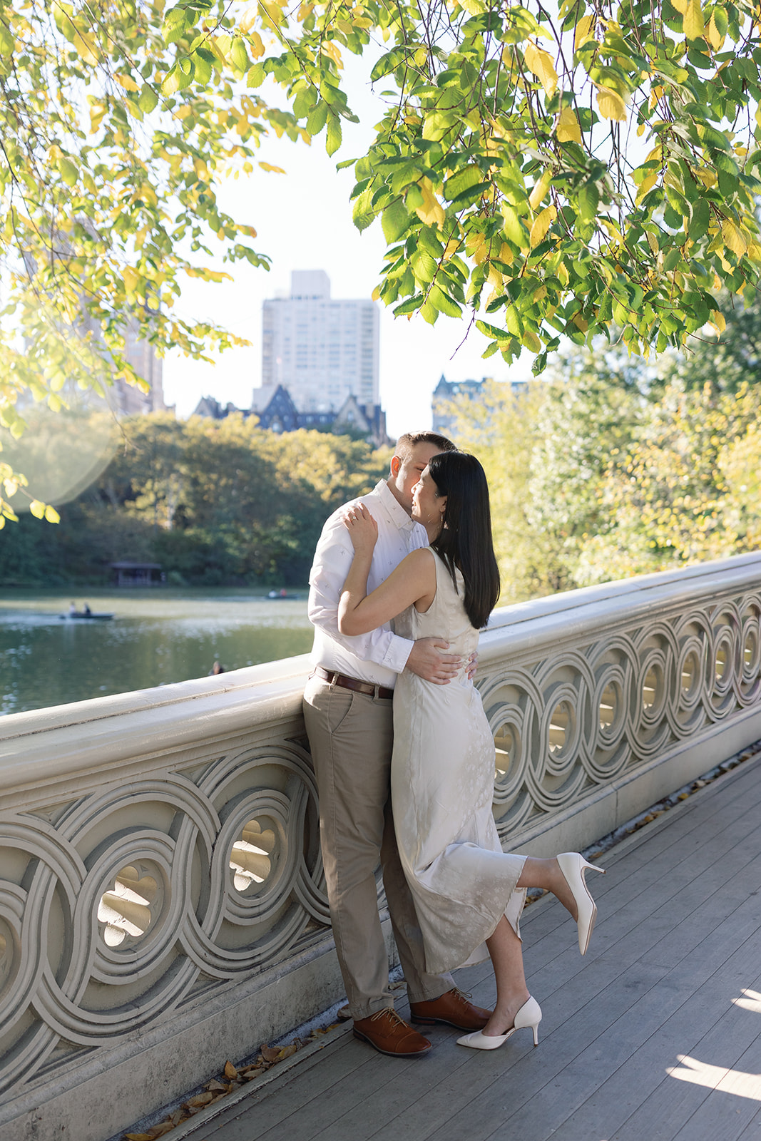 A couple kissing on a bridge with trees and a cityscape in the background, sunlight filtering through leaves.