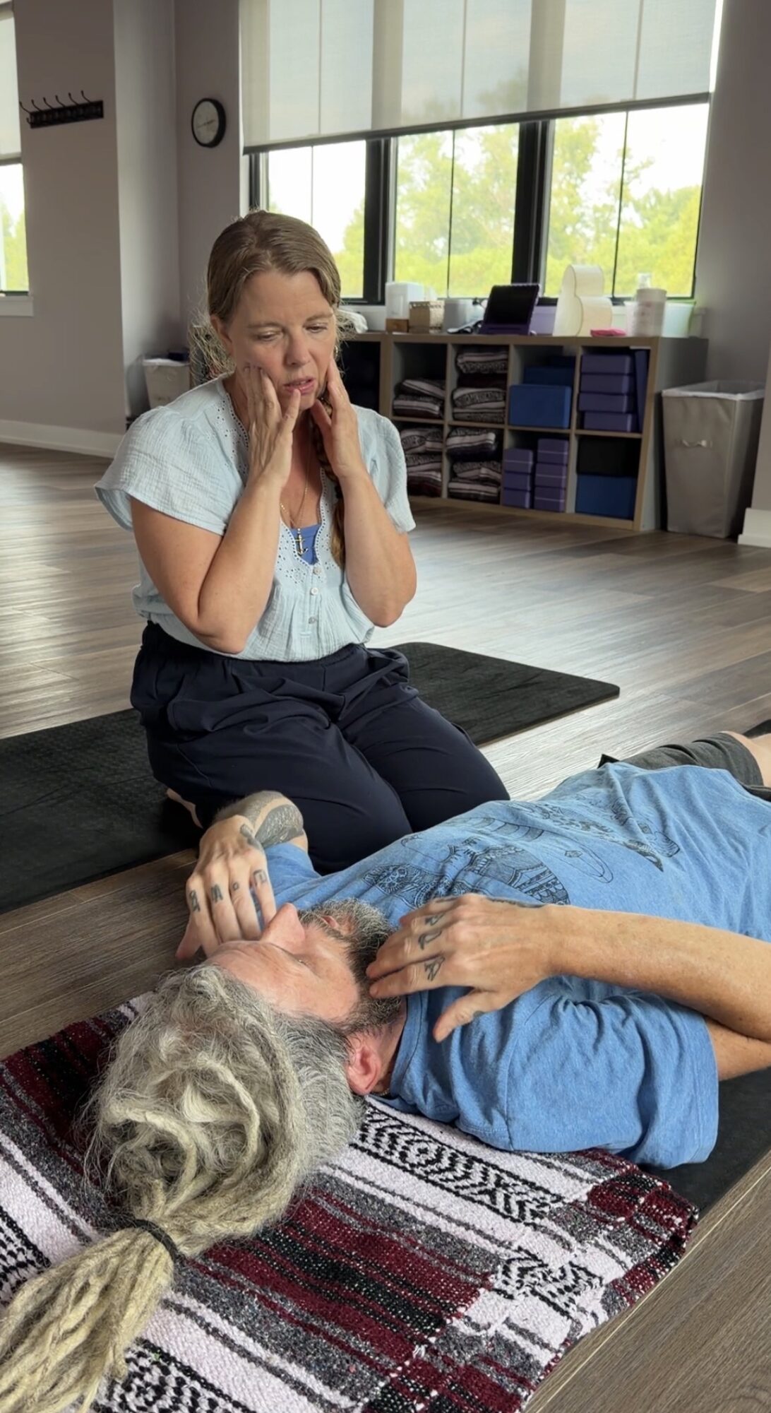 Young girl with hands near face sitting on floor next to an older person lying down, holding their hand, in a room with large windows.