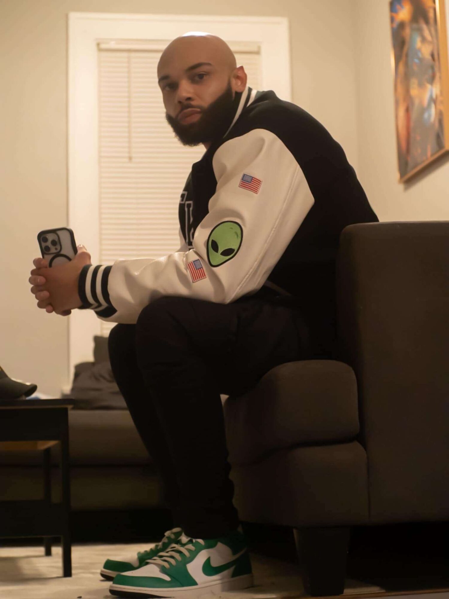 Man with a beard sitting on a couch, holding a phone, wearing a black and white jacket with patches, green and white sneakers.