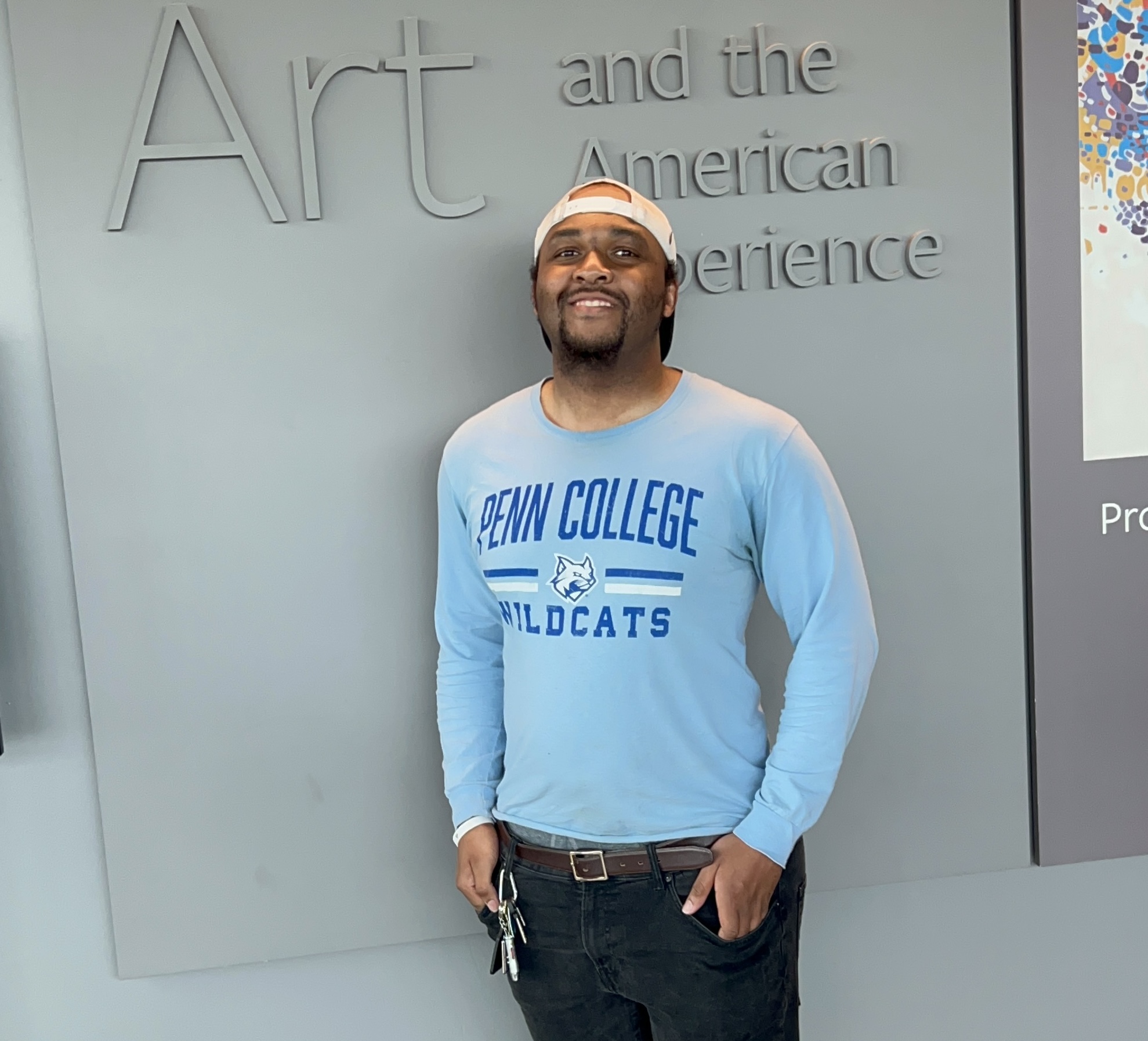 Man wearing a Penn College Wildcats sweatshirt and a white cap, standing in front of a gray wall with text.