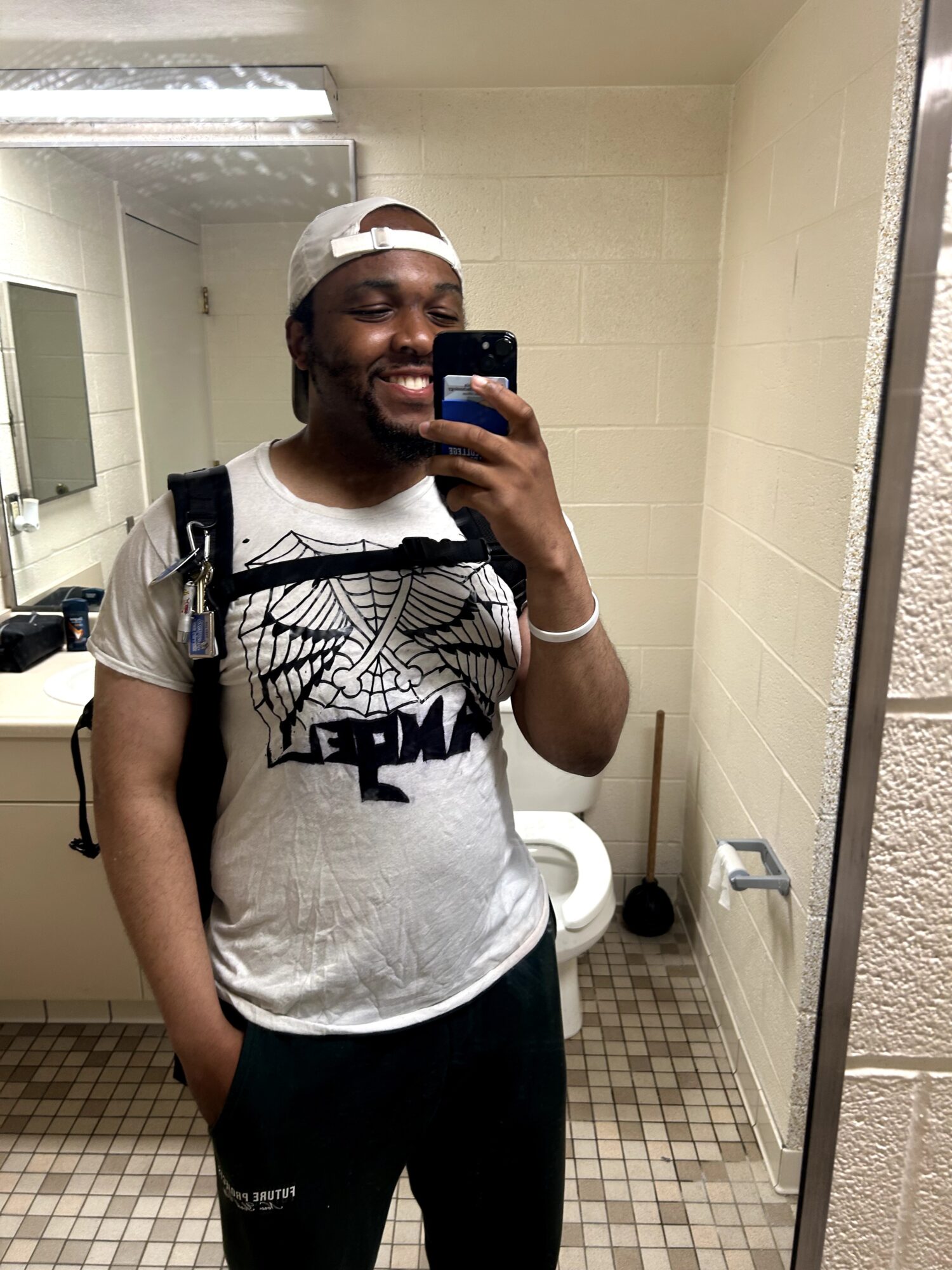 Person taking a mirror selfie in a bathroom, smiling, wearing a white cap, graphic t-shirt, and backpack.
