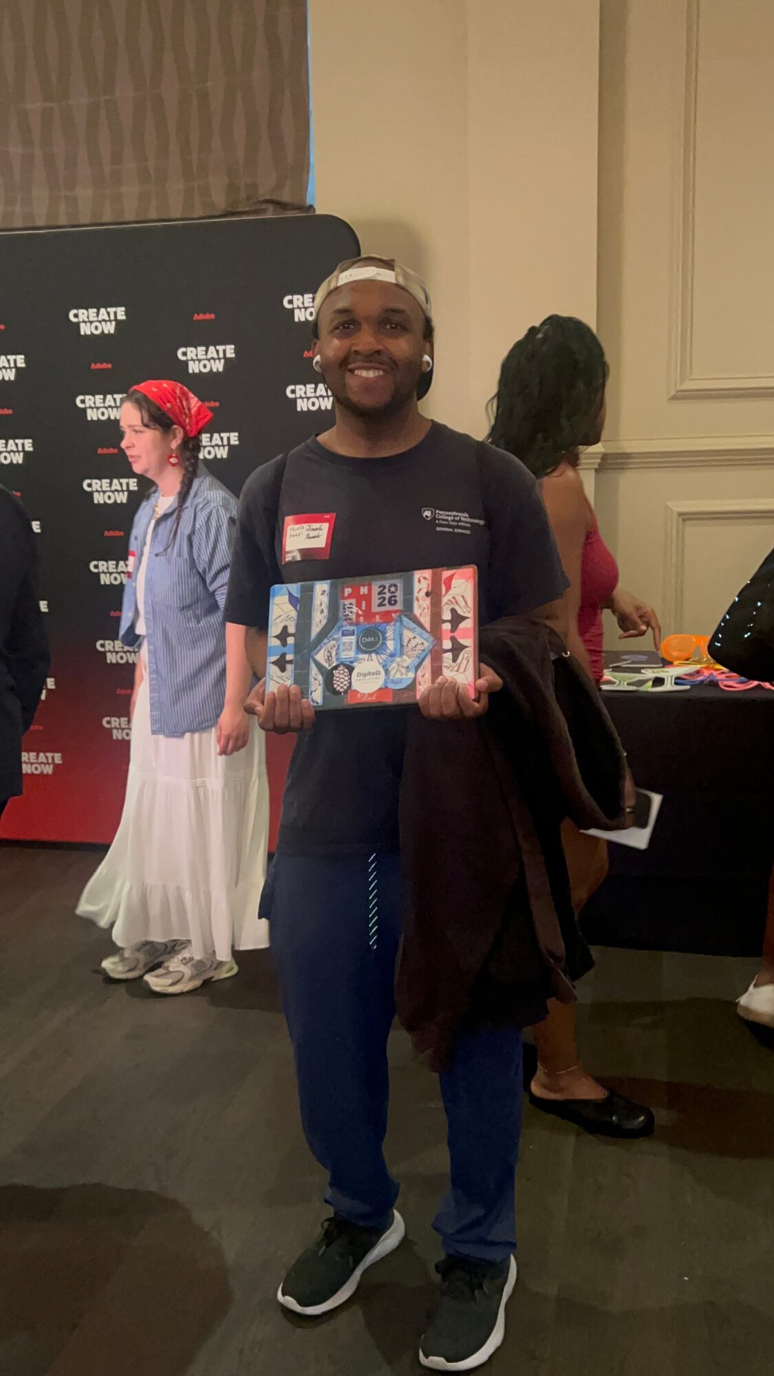 Smiling man holding a colorful gift box at an indoor event, with people and a red backdrop in the background.