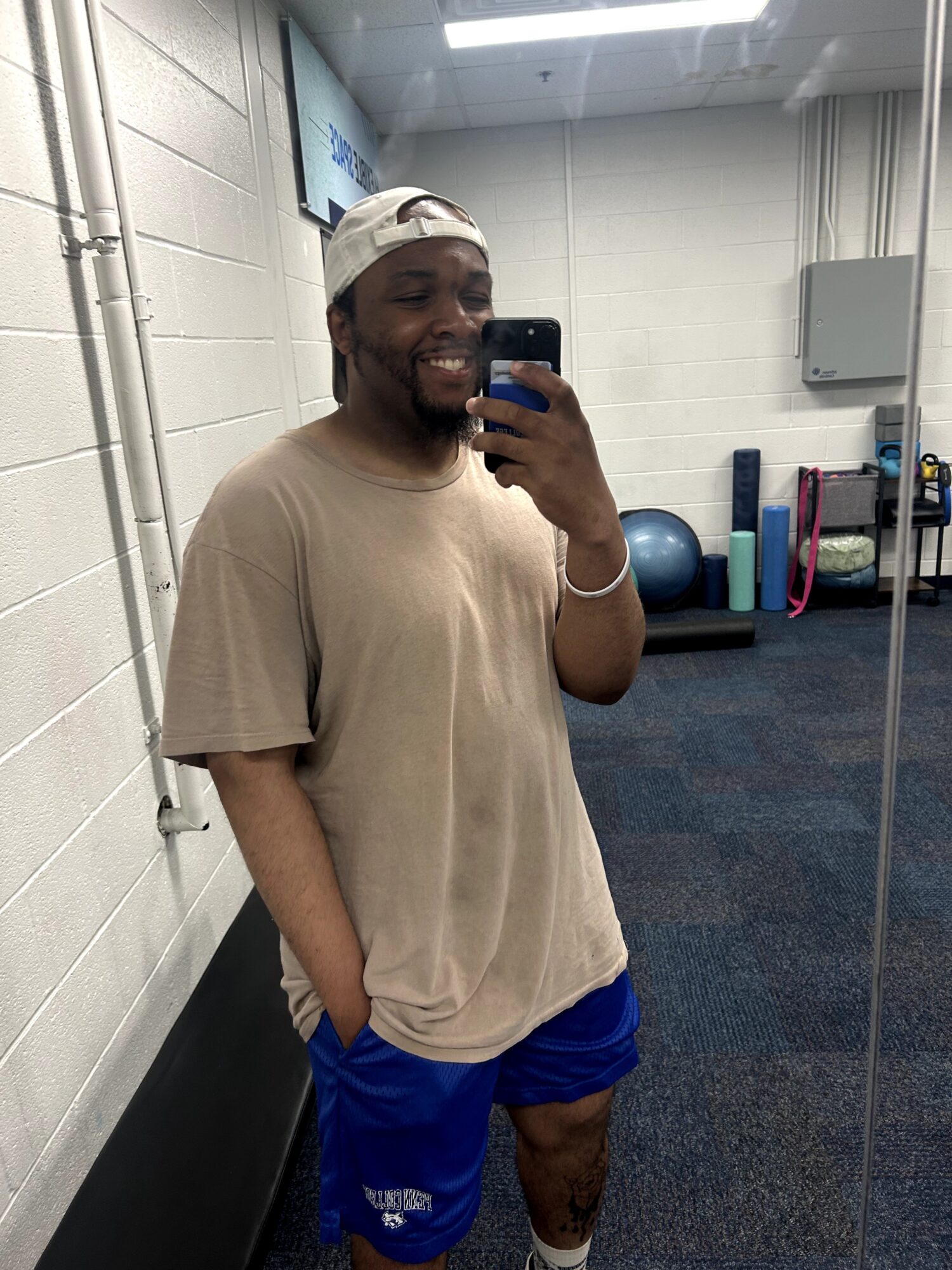 Person taking a mirror selfie in a gym, smiling, wearing a beige t-shirt, blue shorts, and a white cap, with gym equipment in background.