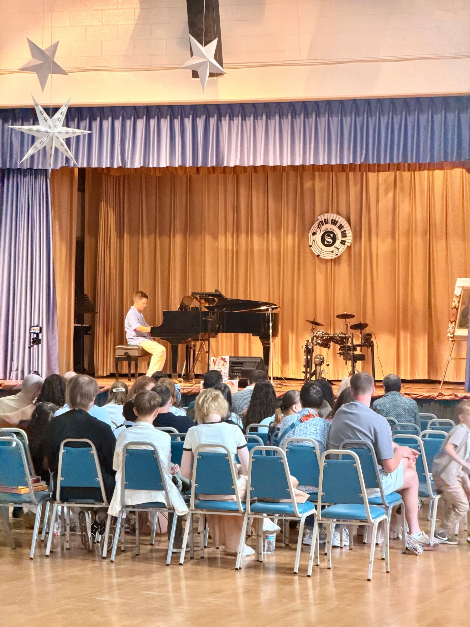 A pianist performs on stage with a drum set and a large curtain, audience seated in chairs watching the performance.