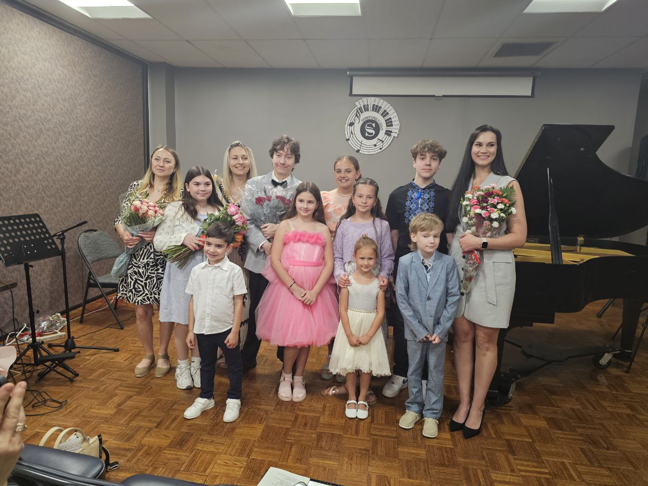 Group of children and two adults standing in a music room with a piano, holding flowers, smiling, with a logo on the wall.