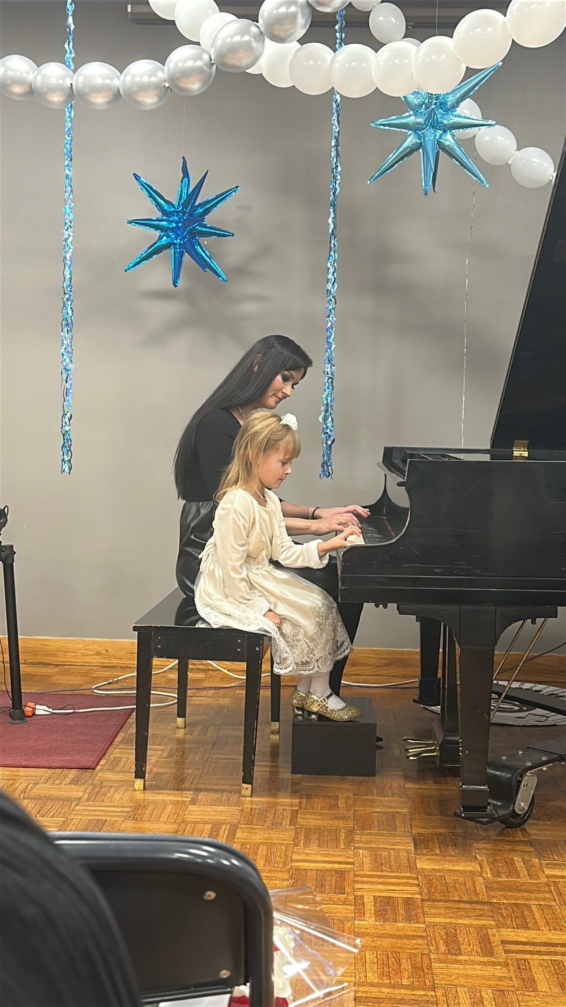 A woman and a young girl play piano on stage decorated with blue and white star and bead ornaments.