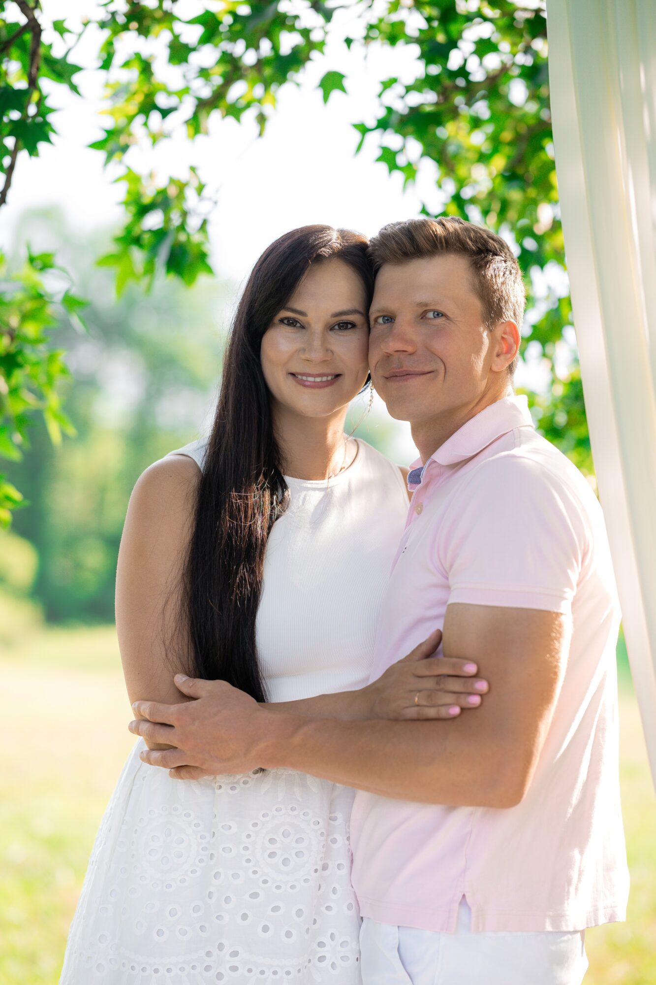 A smiling couple stands outdoors with greenery in the background, embracing each other, woman in white and man in pink shirt.