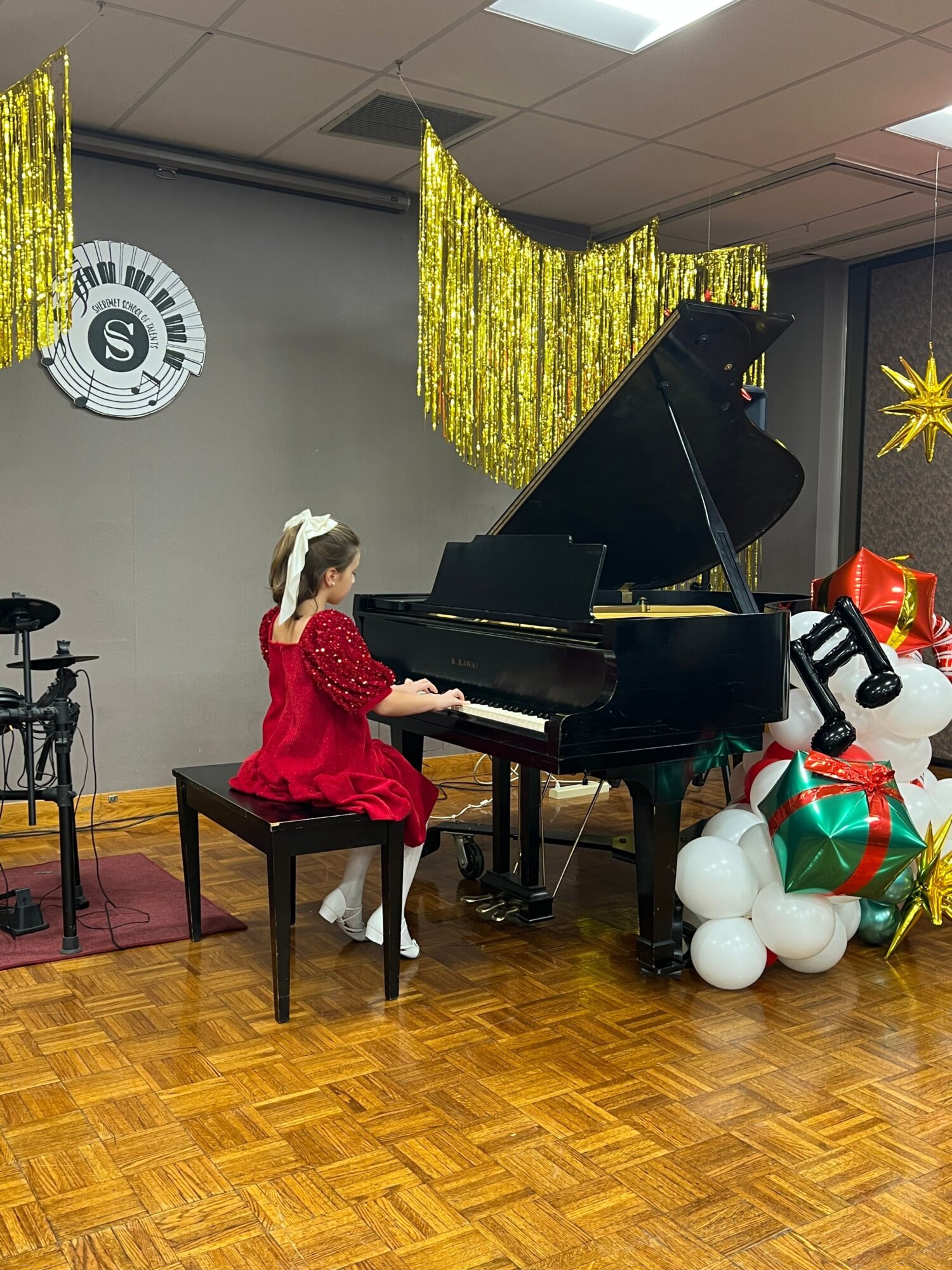 Child in red dress sitting at a black piano decorated with Christmas balloons and ornaments, on a wooden floor.