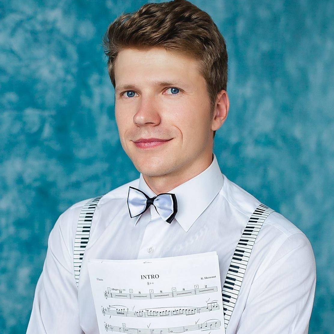 Young man with short brown hair, wearing a white shirt, black bow tie, and suspenders, holding sheet music, smiling, blue background.