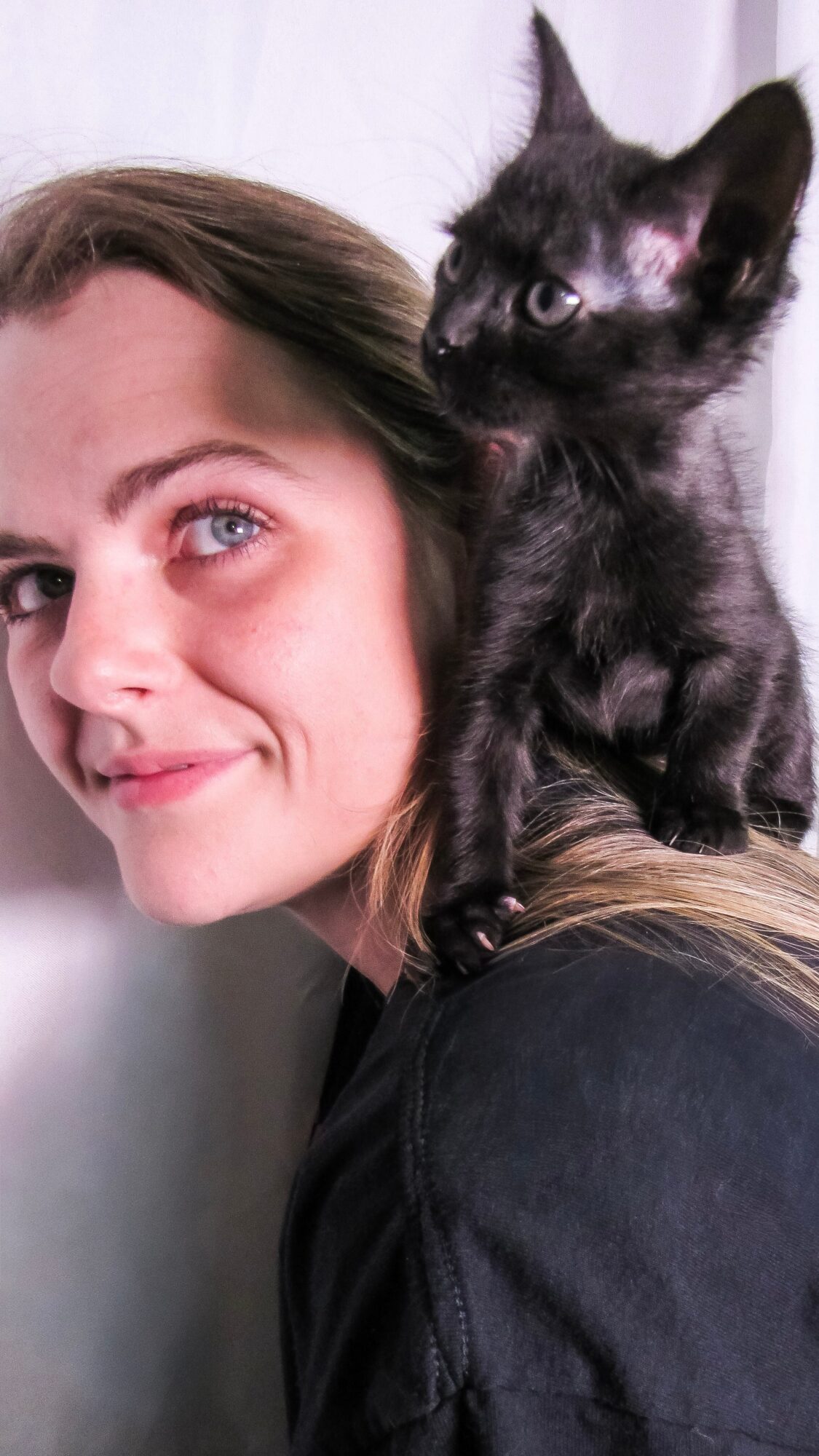 Woman with long hair and blue eyes, smiling, with a black kitten on her shoulder, against a light background.