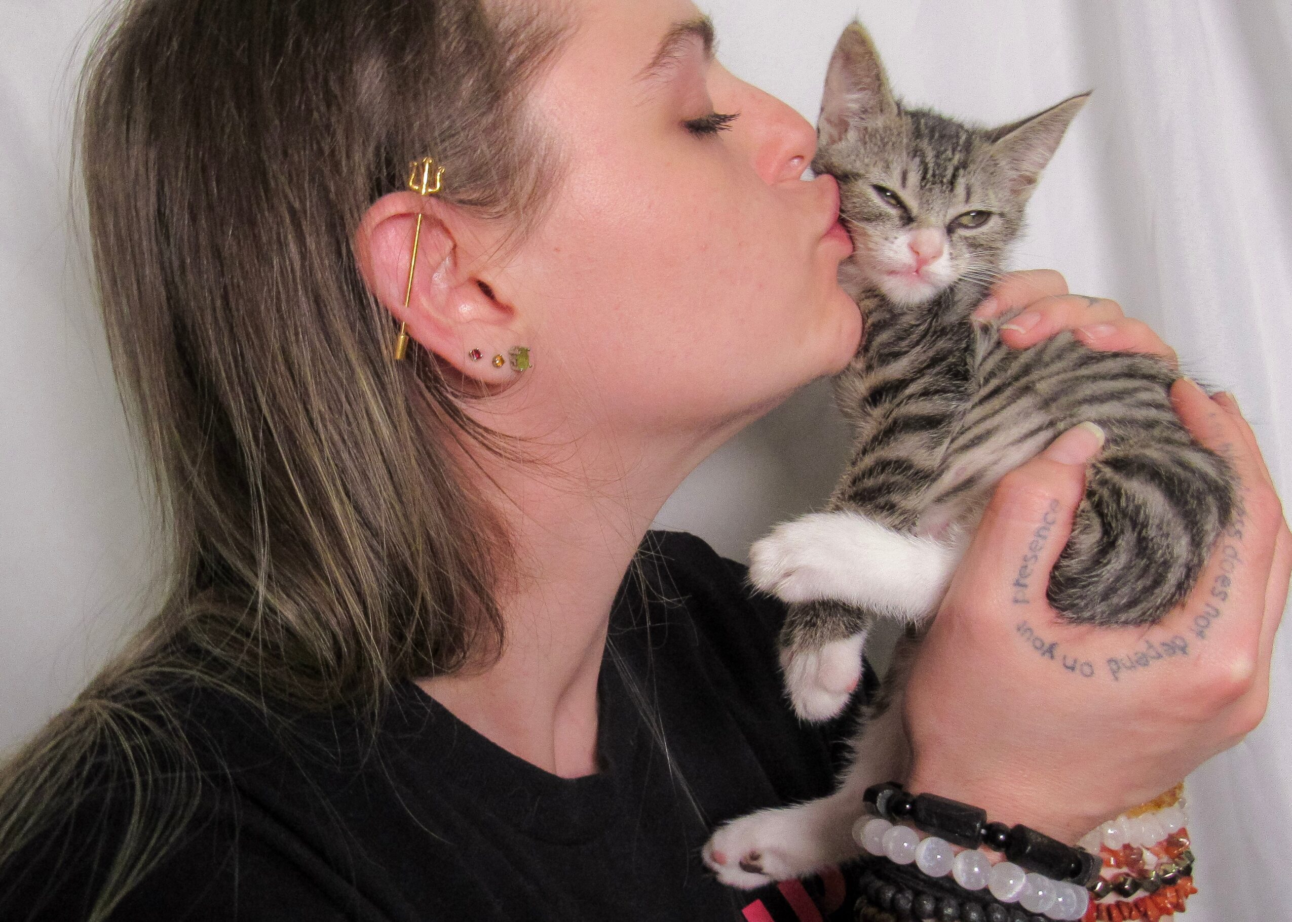 Person holding a striped kitten close to face, about to kiss it, with a white curtain background.
