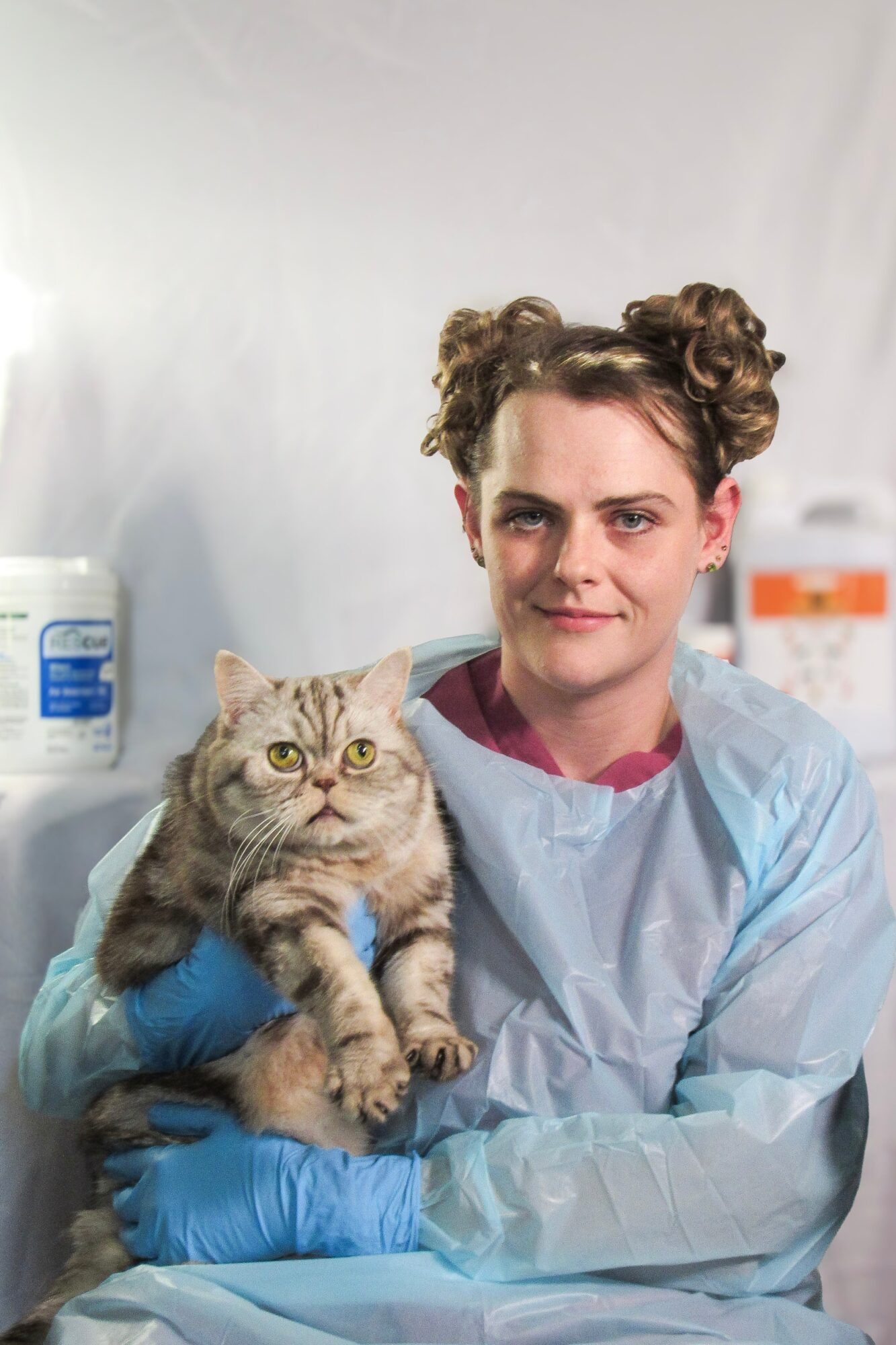 Young woman in medical gown holding a tabby cat, both wearing blue gloves, in a clinical setting.