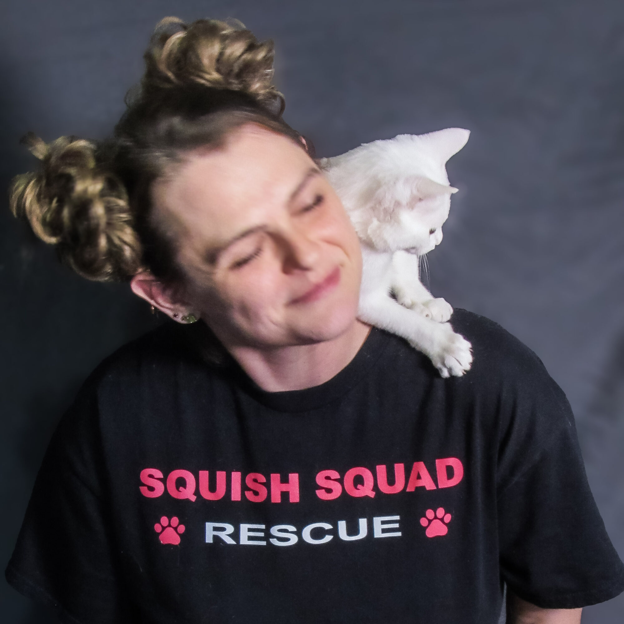 Person with a cat on shoulder, smiling with eyes closed, wearing a black T-shirt with pink and white text, against a dark background.
