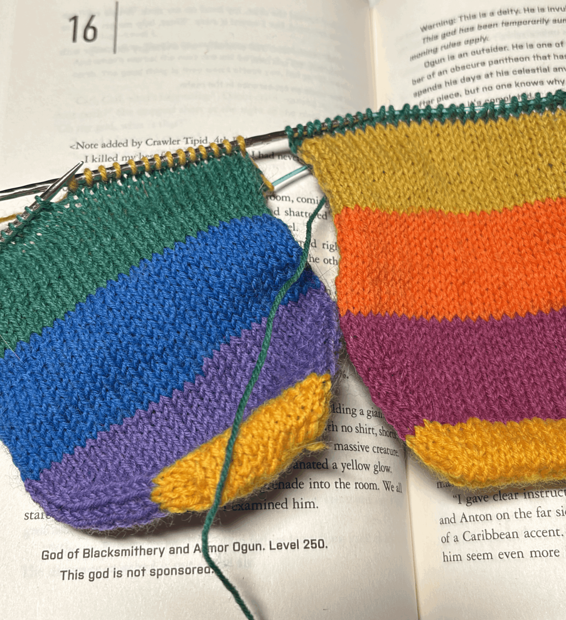 Two knitted pieces with rainbow stripes on an open book, knitting needles inserted, yarn attached, colorful and textured.