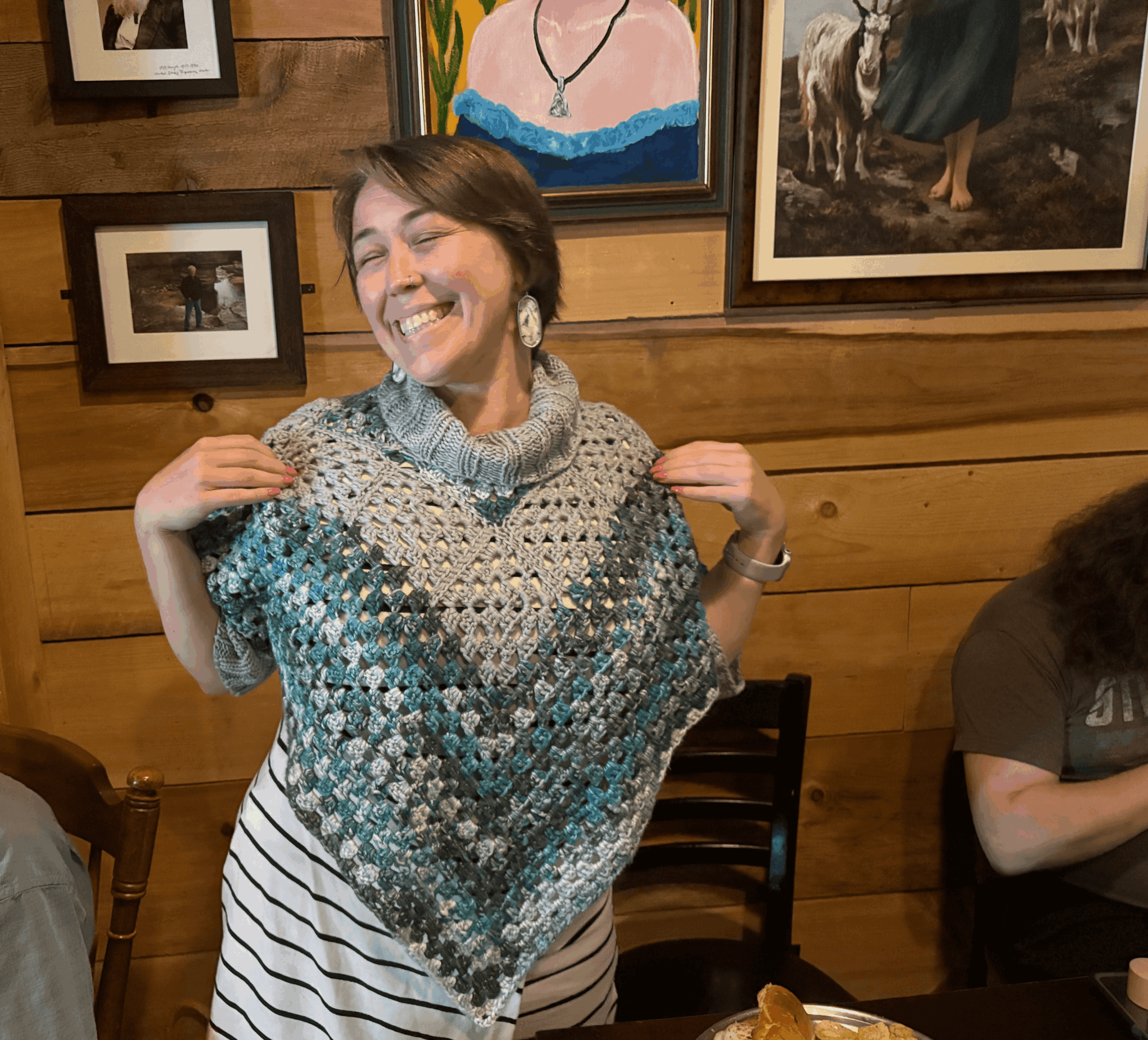 Woman smiling and holding a colorful shawl in a wooden-paneled room with framed artwork on the wall.
