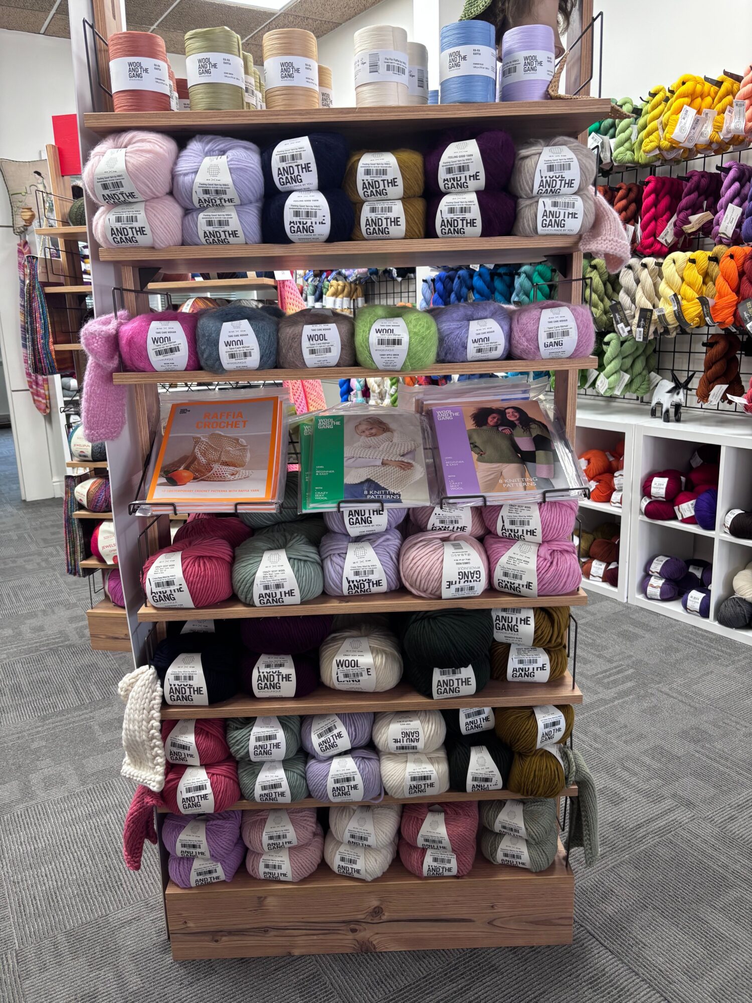Display of yarn skeins in various colors on wooden shelves, with knitting supplies in the background.