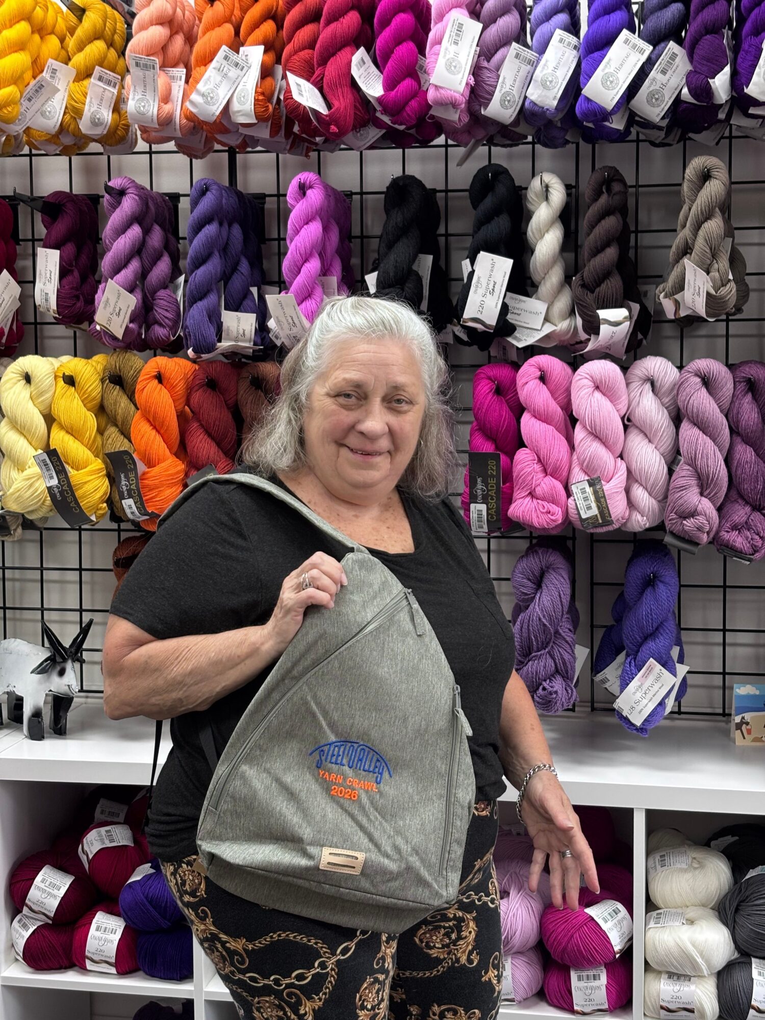 Woman shopping for yarn in a store, holding a gray bag, surrounded by colorful yarn skeins on shelves.