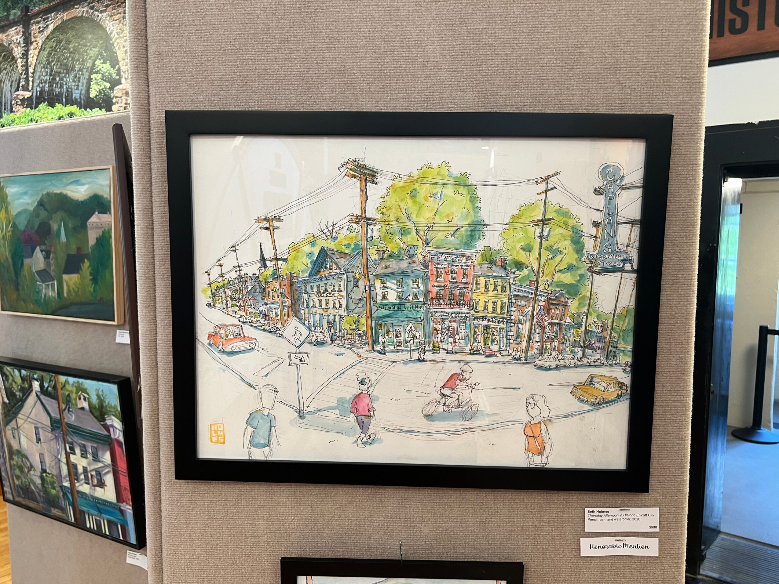 Colorful street scene with people, cars, trees, and buildings, framed and displayed on a wall.