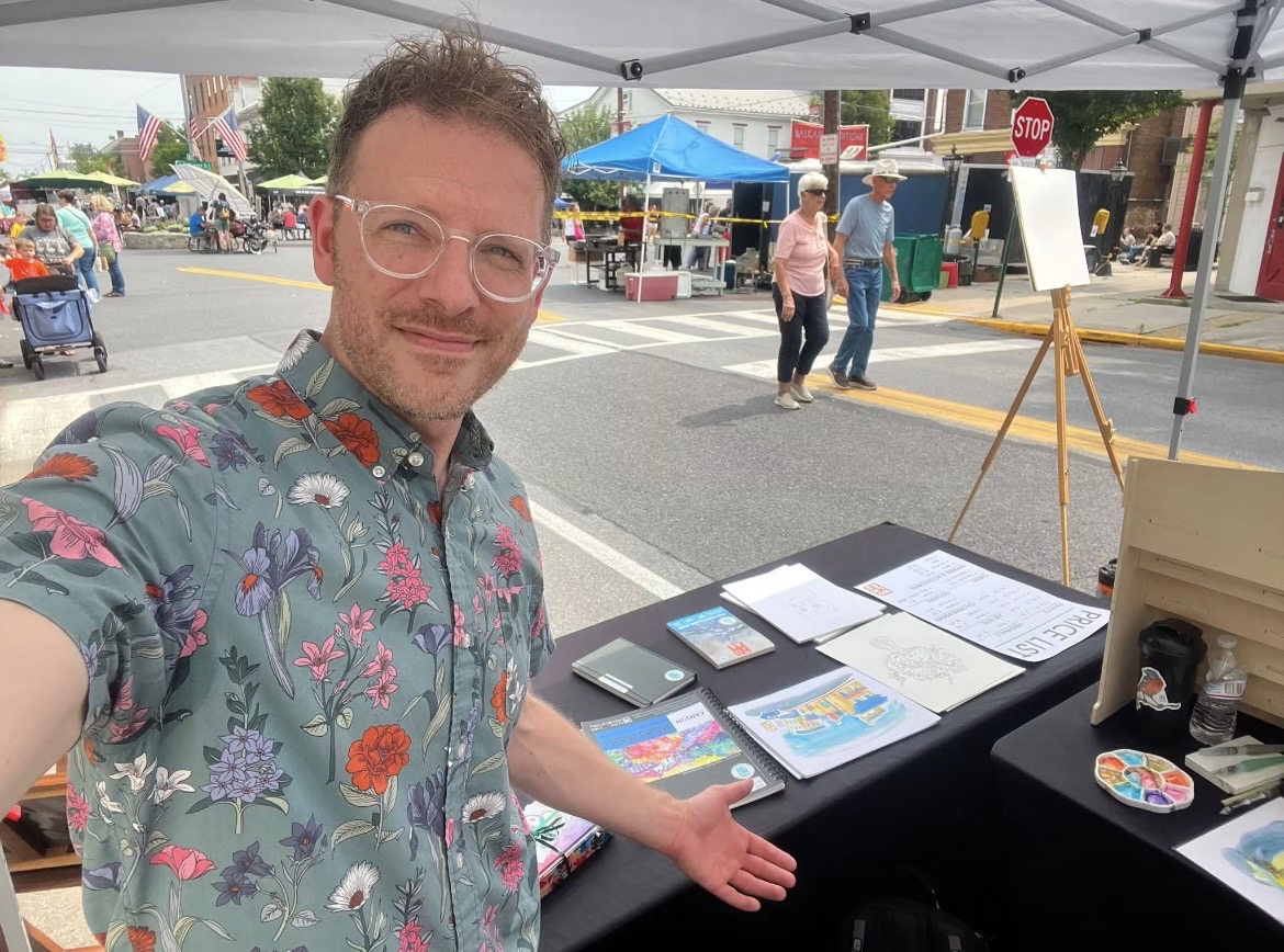 Man with glasses in a floral shirt at outdoor market booth, smiling, with tables and people in background.