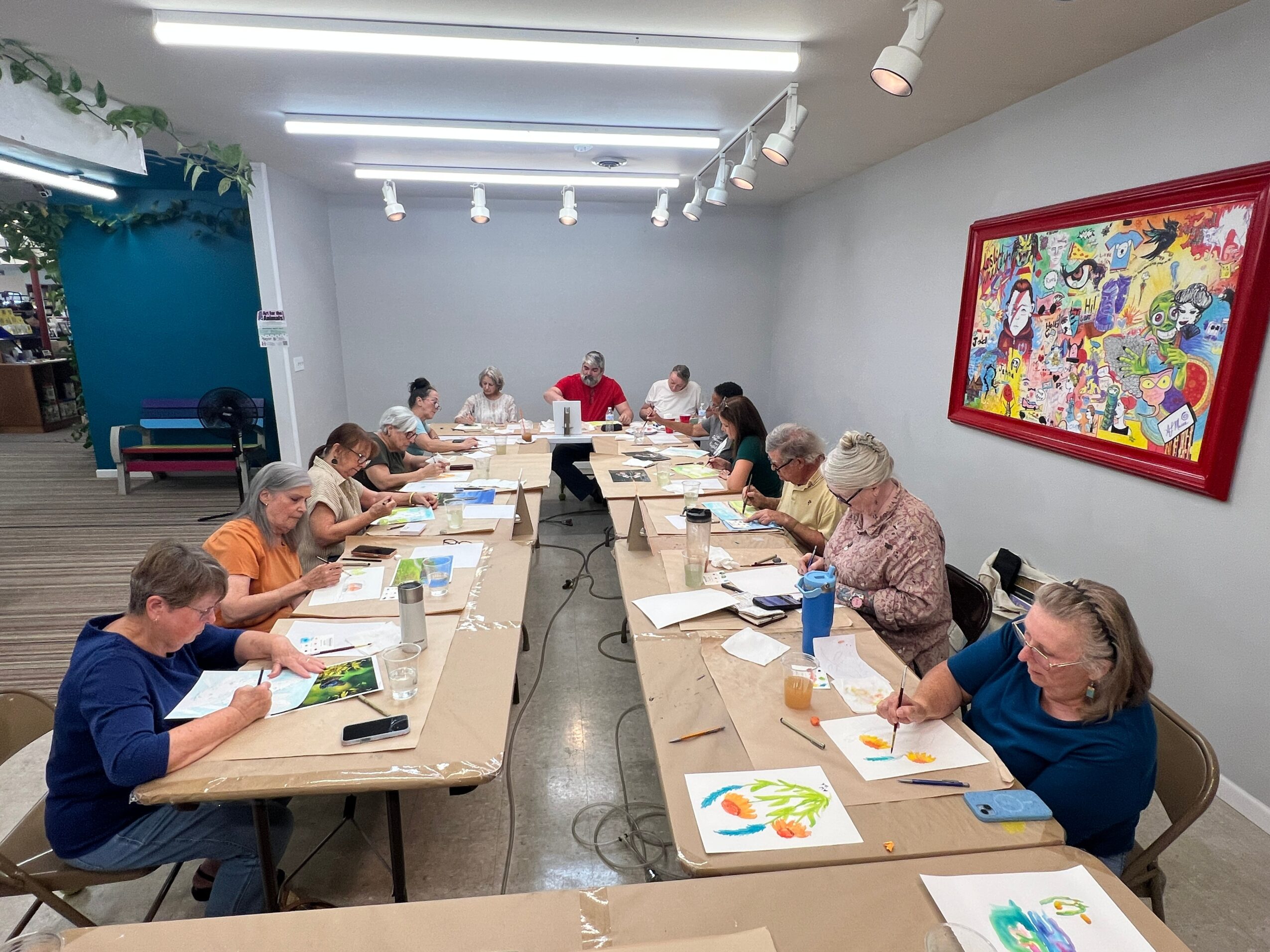 Group of people sitting around a long table in a room, engaging in a creative activity, with colorful artwork on the wall.