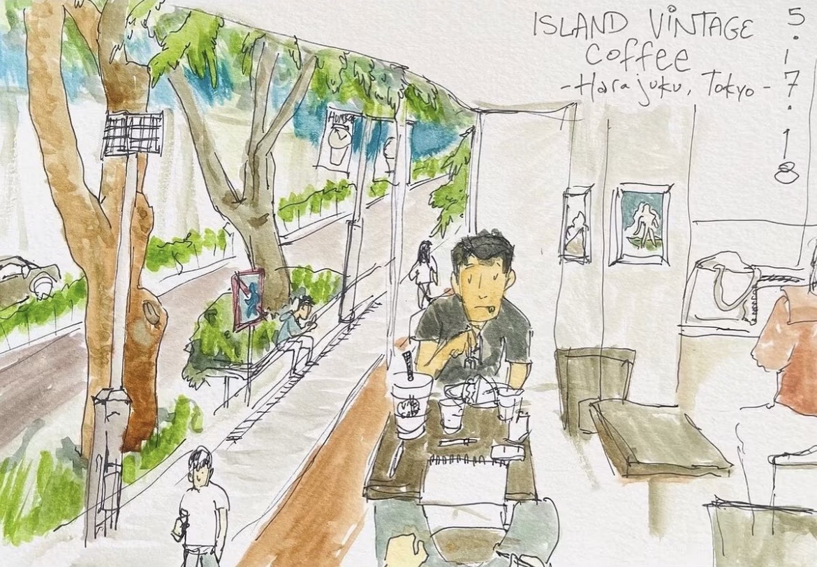 Man sitting at a table in a cafe with trees outside, people walking, and framed pictures on the wall.