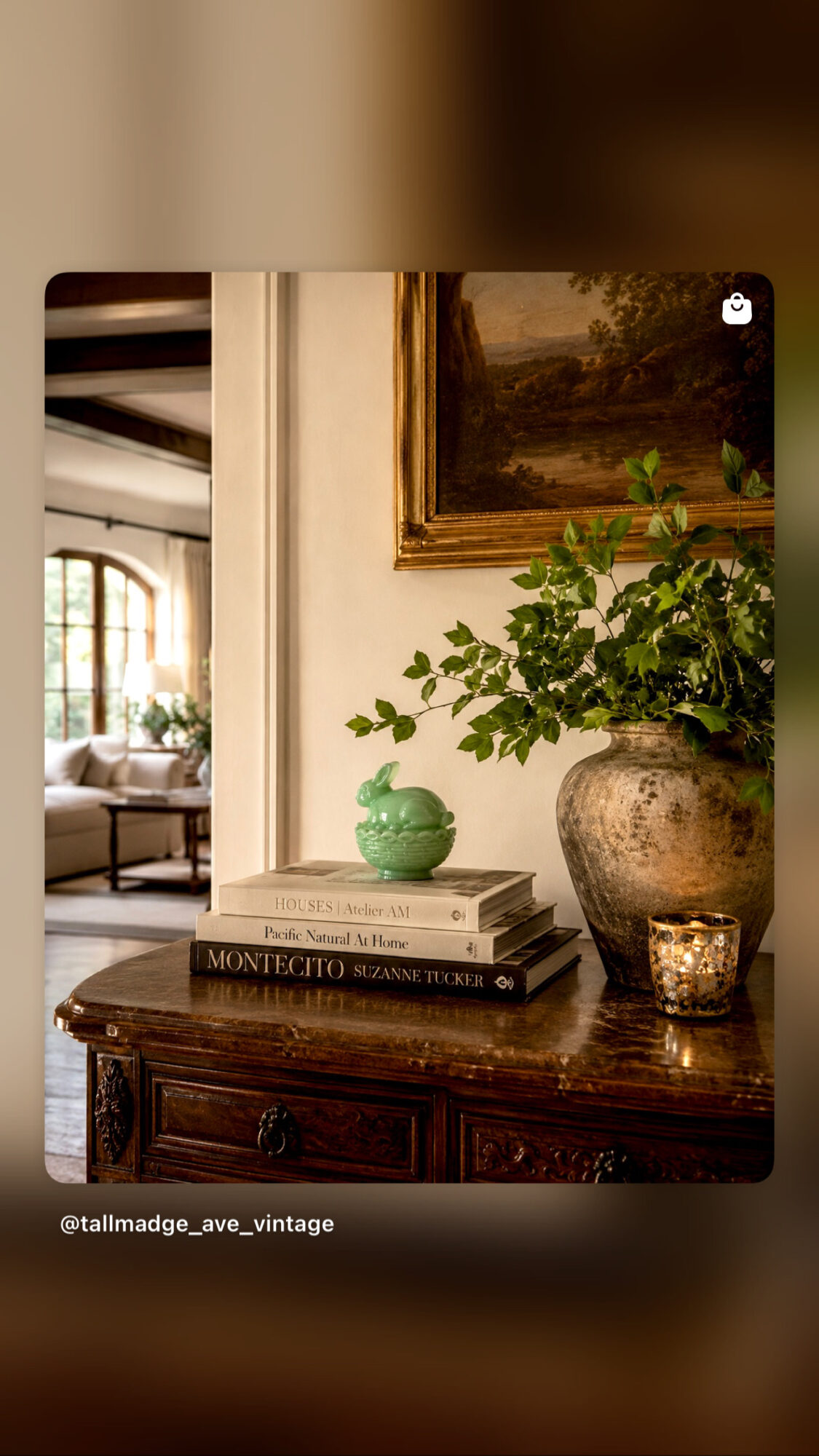 A wooden table with a stack of books, a green vase, a large potted plant, and a candle in a glass holder, in a room with a painting and window.