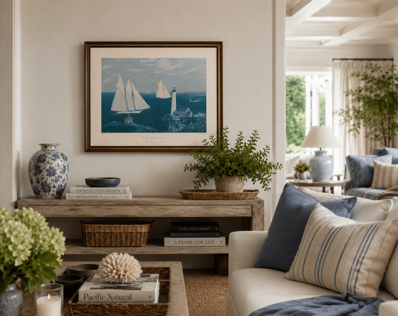 Living room with framed nautical painting, potted plant, and decorative pillows on a sofa, bright window in background.
