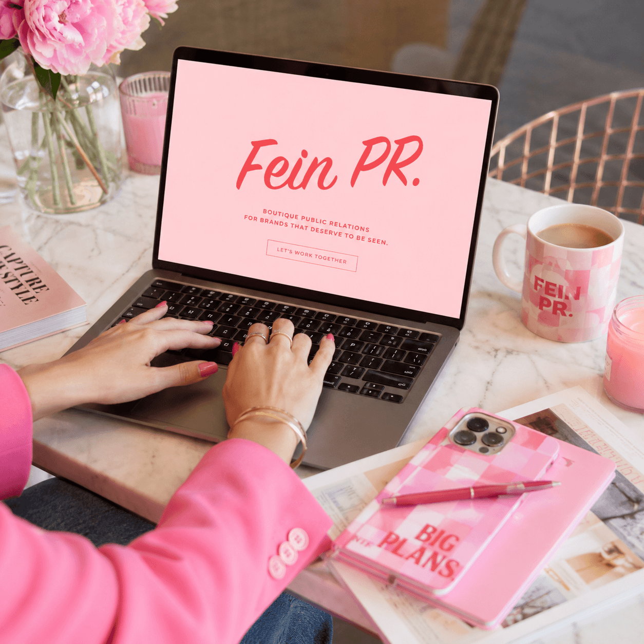 Person in pink jacket typing on laptop with pink notebook, pen, coffee mug, and flowers on table.