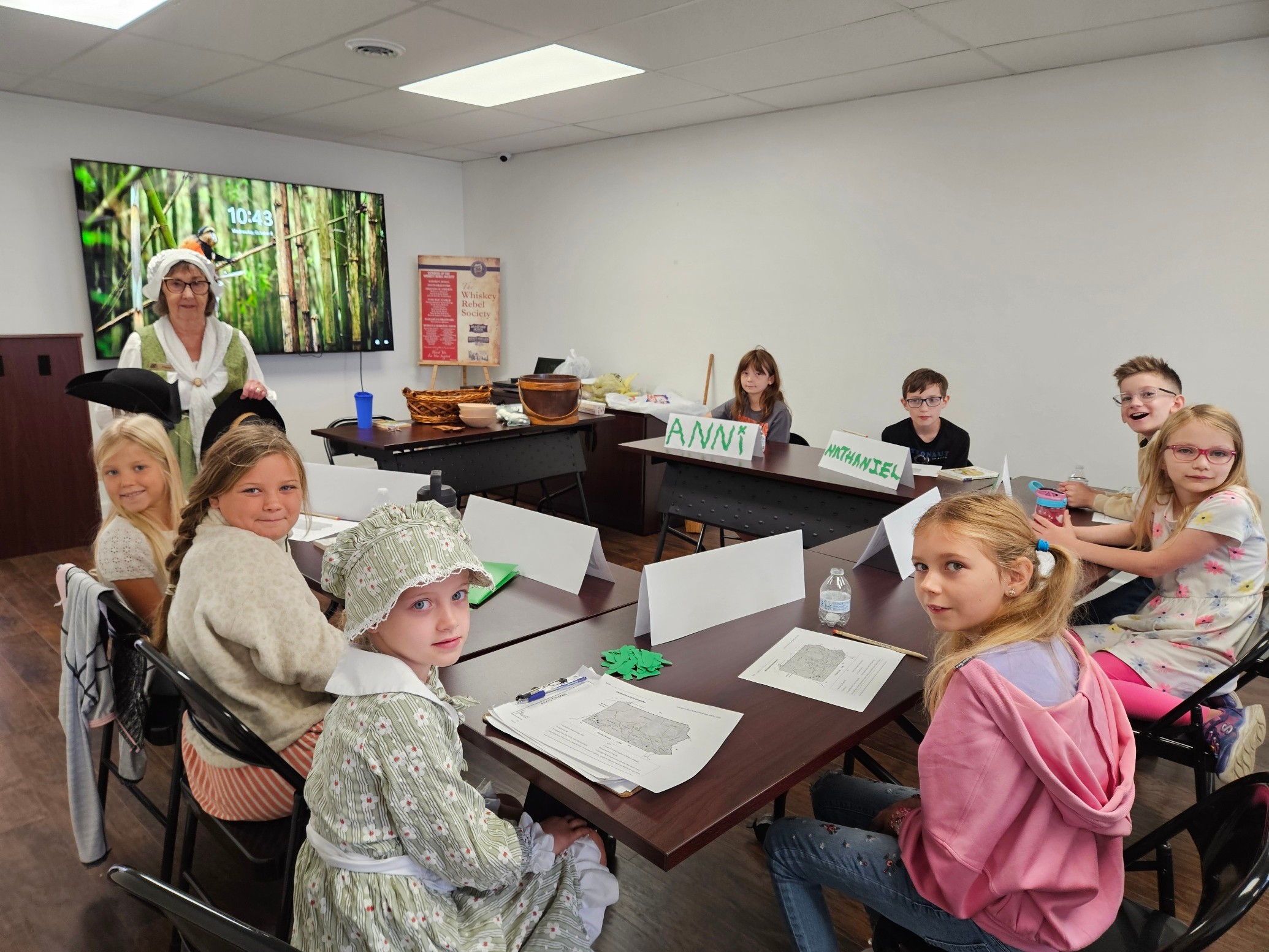 Children sitting at tables in a classroom, with a teacher on a screen on the wall, some with notebooks and supplies.